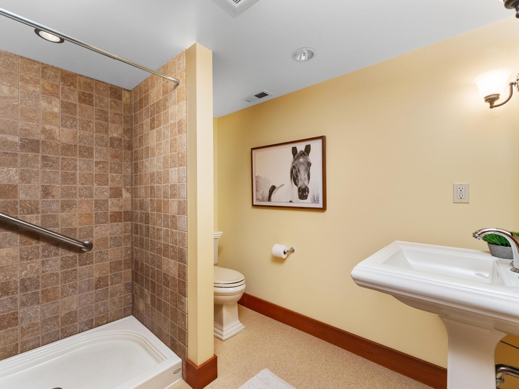 A cozy Portland, Oregon bathroom features a tiled shower, white pedestal sink, and toilet. The yellow walls add warmth, with a framed horse picture above the toilet and a sconce light beside the mirror for perfect illumination.
