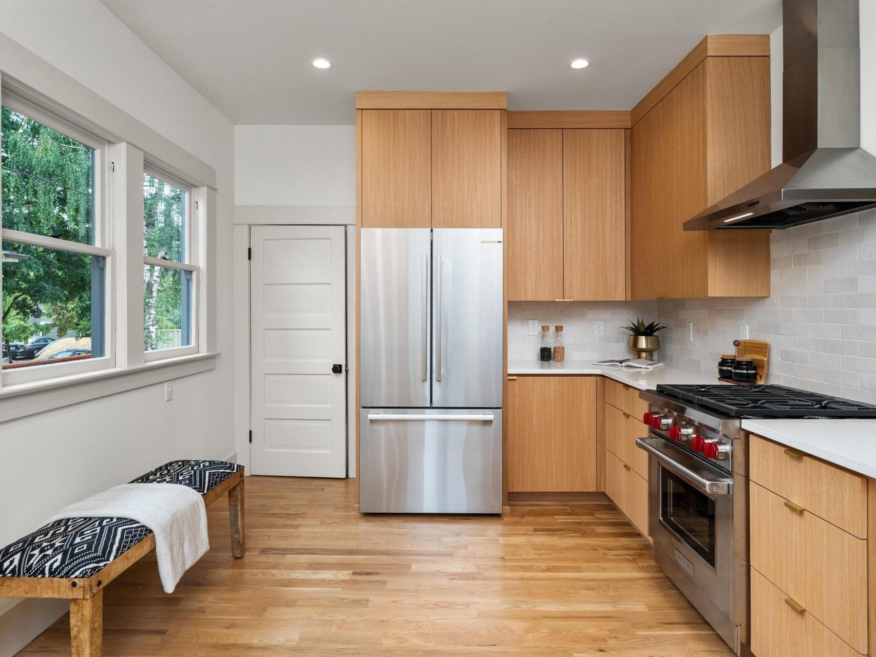A modern kitchen in Portland, Oregon, showcases wooden cabinets and hardwood floors with a stainless steel refrigerator and stove. The white tiled backsplash contrasts elegantly with the wood. A bench featuring a patterned cushion sits below the windows on the left side.