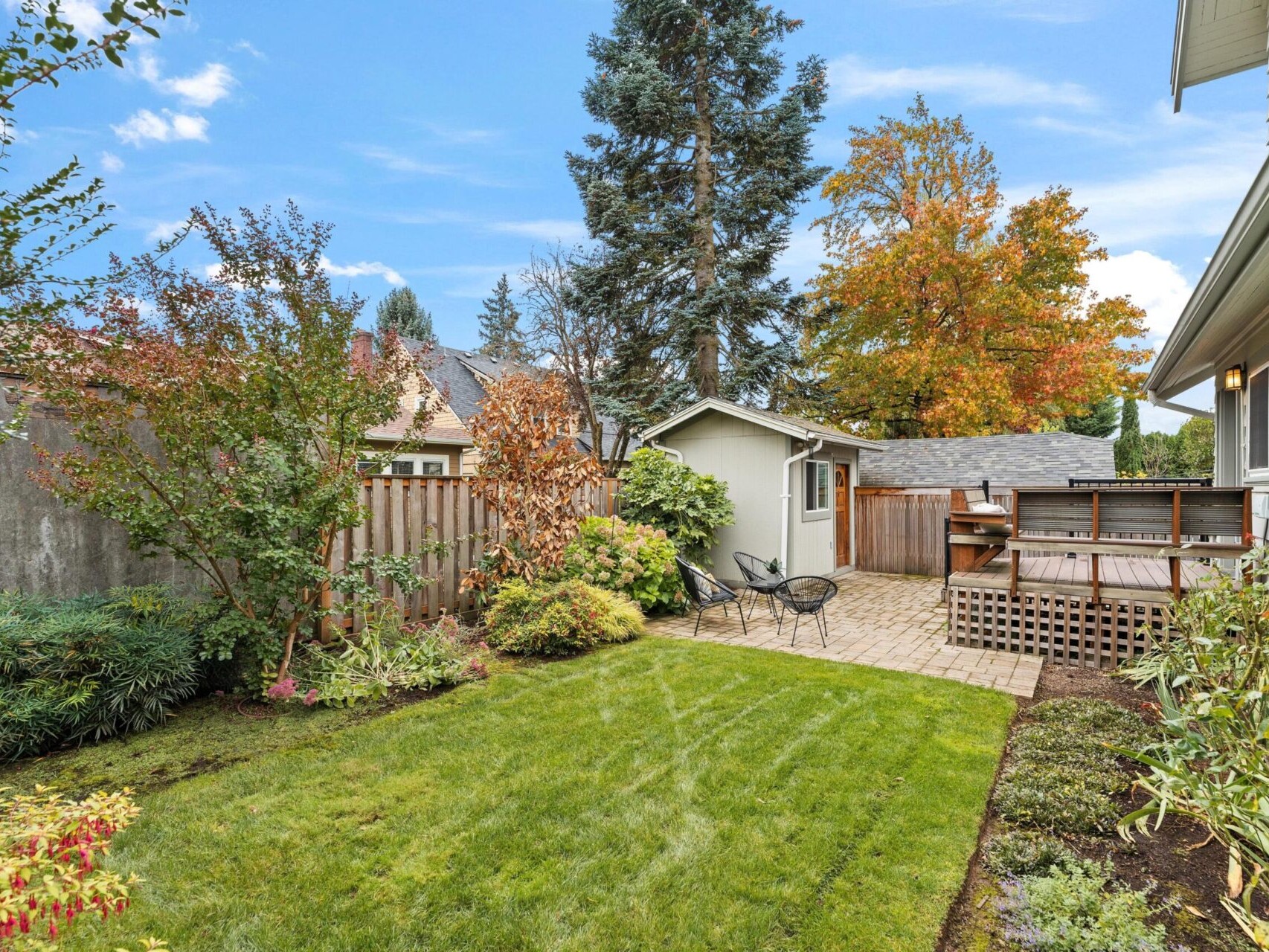 A backyard with a well-manicured lawn, surrounded by trees and plants. A small patio area features a couple of chairs, and a wooden fence encloses the space. A detached shed is in the background, and colorful autumn foliage is visible.