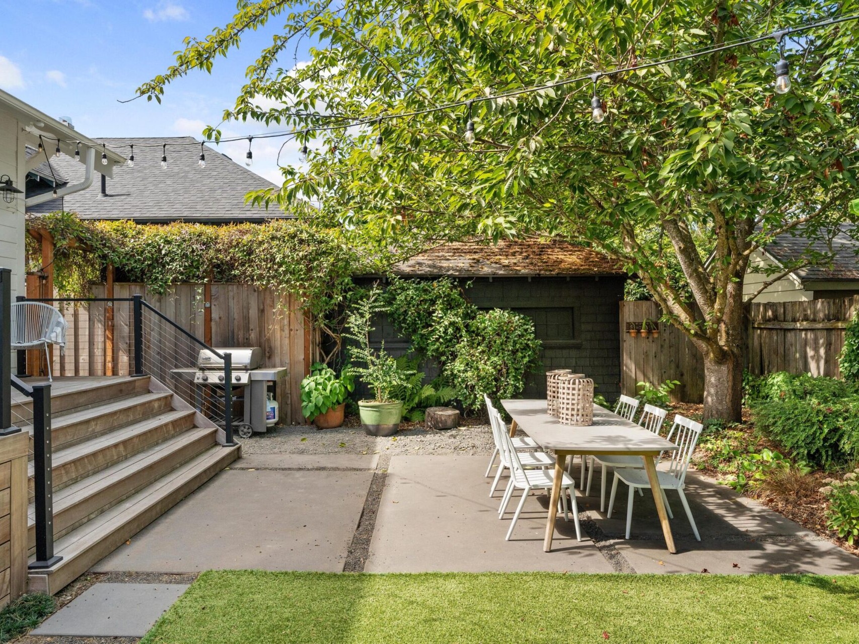 A backyard scene in Portland, Oregon, features a modern patio with a wooden dining table and white chairs under a large tree. Steps lead up to the house with string lights above. A barbecue grill and small shed nestle near the wooden fence, blending charm and functionality.