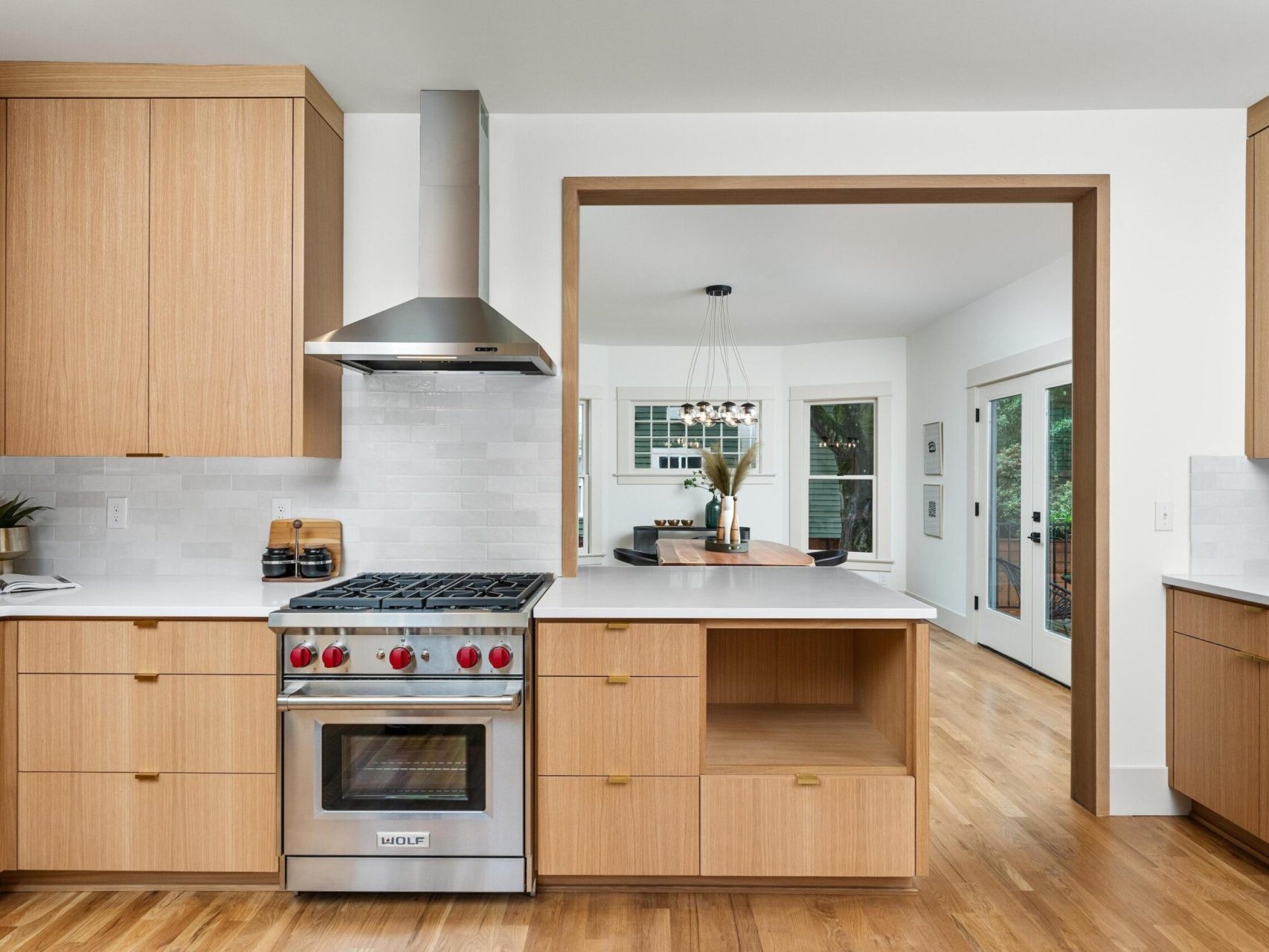 A modern kitchen in Portland, Oregon features wooden cabinets, a stainless steel range with a hood, and a light-colored backsplash. The room opens into a dining area with a table and large windows offering views of the lush greenery outside.