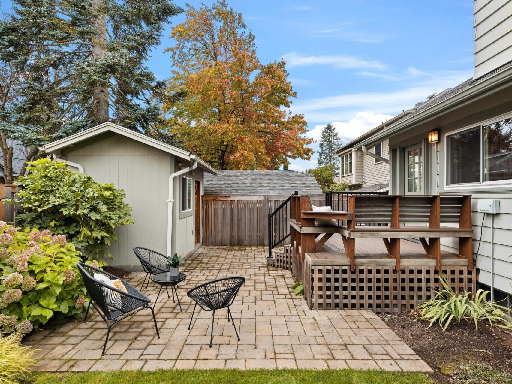 A backyard with a small wooden deck attached to a house. There are three black metal chairs on a brick patio. A shed is nearby, surrounded by greenery and a wooden fence. Trees with autumn foliage are in the background under a clear blue sky.