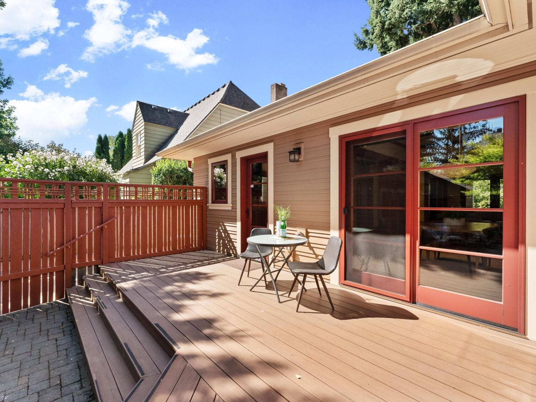 A sunny patio with a wooden deck in Portland, Oregon, features a small table and two chairs. The house boasts red-framed glass doors and a light beige exterior. A wooden fence and trees stand in the background under a blue sky with scattered clouds.