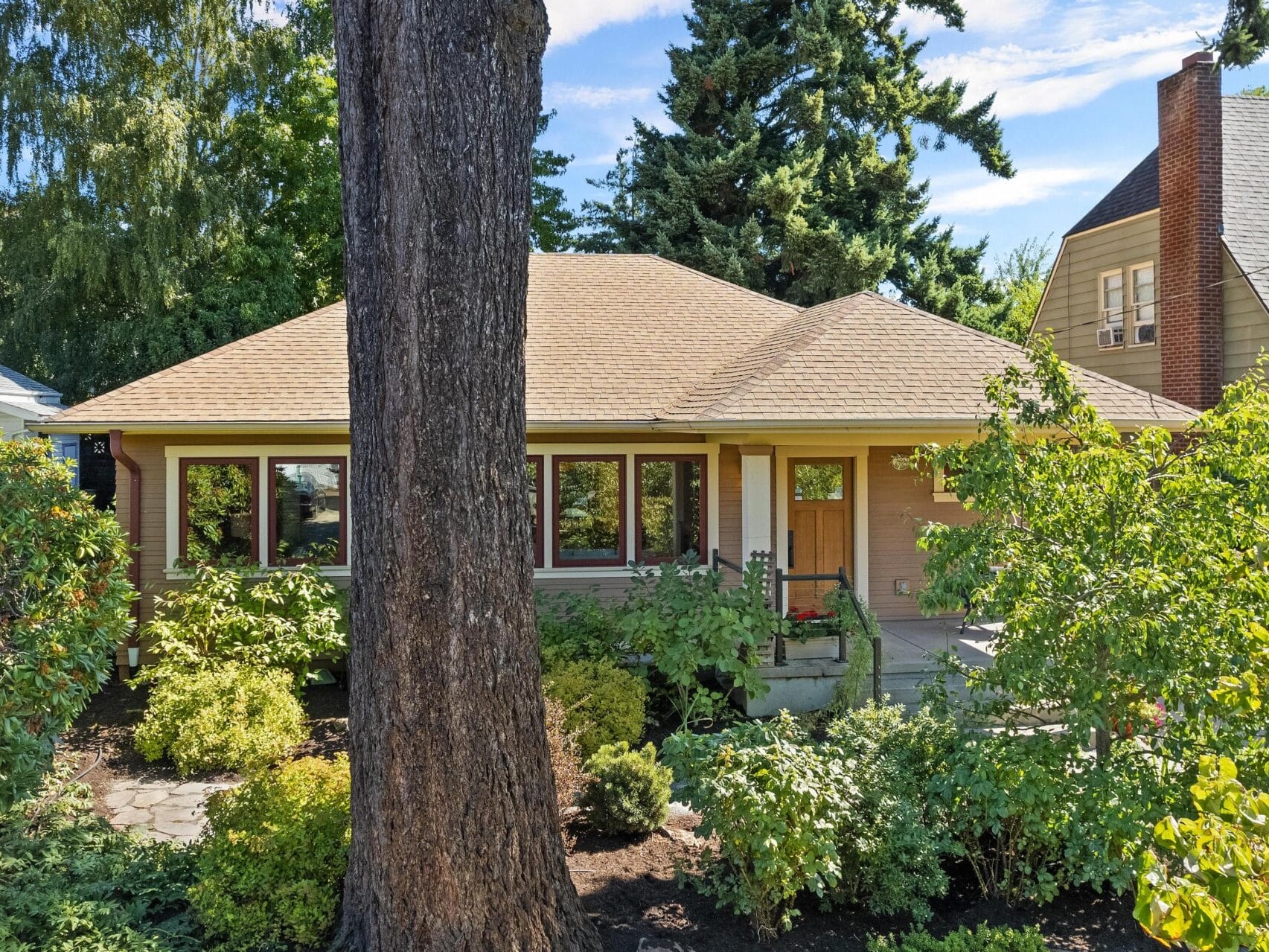 A small, charming house with a light brown exterior and a covered porch, partially hidden by a large tree. Nestled in the lush greenery and shrubs of Portland, Oregon's residential neighborhoods, it basks under the sunny sky.
