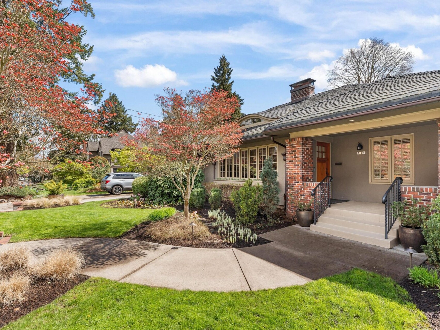 A charming suburban house with a brick facade is nestled in Portland, Oregon. Surrounded by a well-maintained garden, it boasts a paved walkway, lush green lawn, and red-leafed trees. A car rests in the driveway under a clear blue sky, awaiting its new owner through your top Portland realtor.