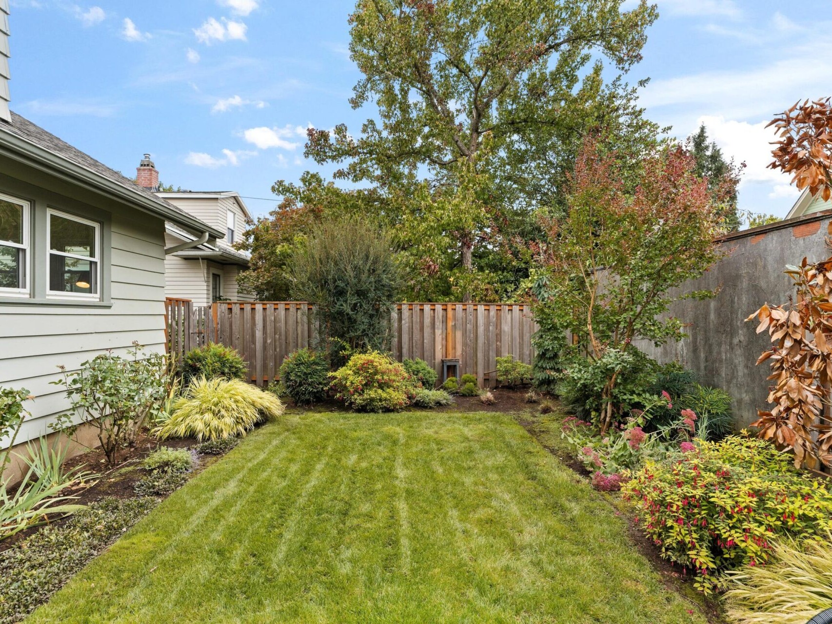 A well-maintained backyard featuring a neatly mowed grassy lawn. Its bordered by various shrubs and colorful plants, surrounded by a wooden fence. A tall tree and a house with green siding are visible in the background under a clear blue sky.