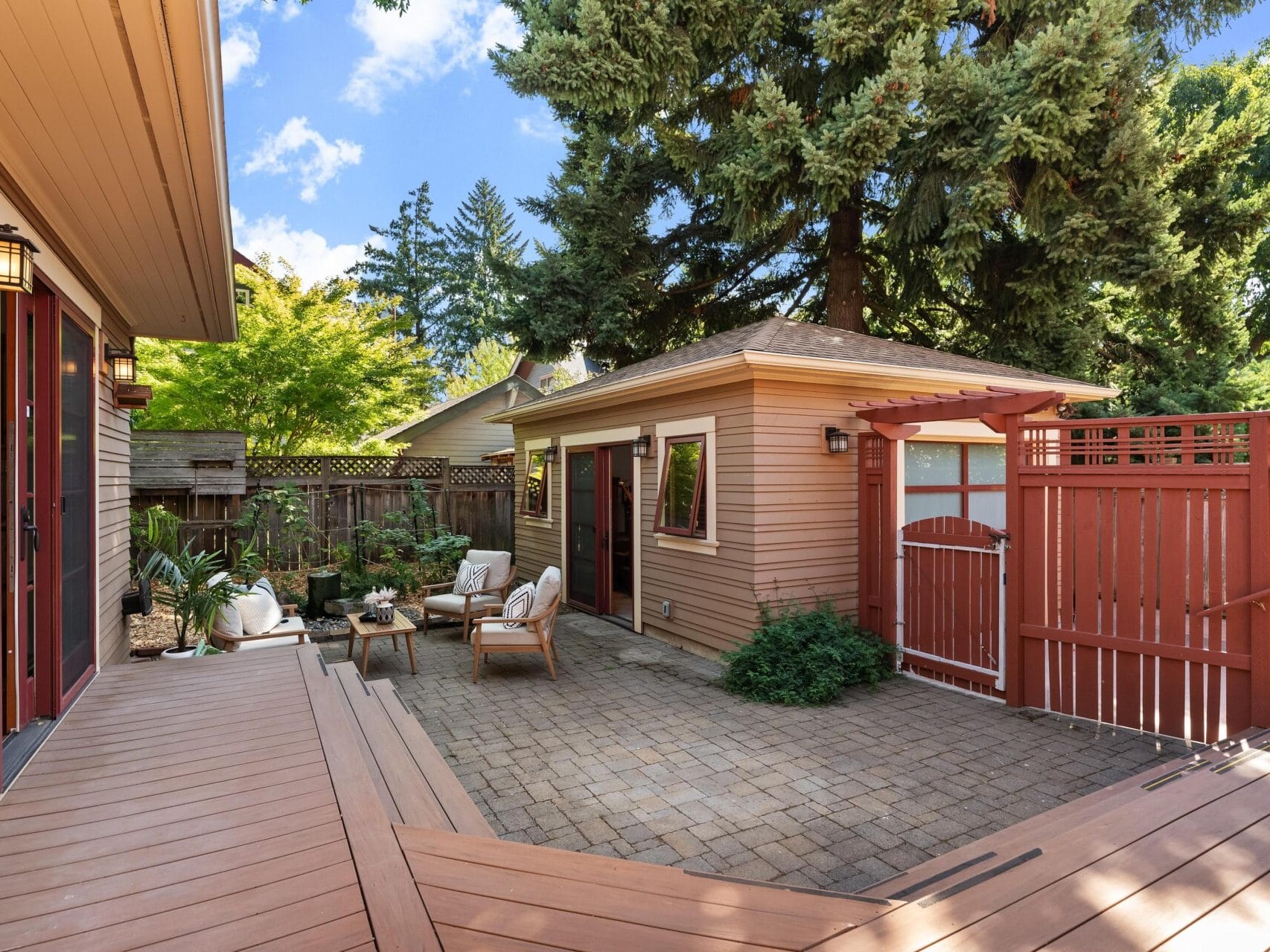 A cozy Portland, Oregon backyard features a small seating area with chairs and a table, surrounded by trees and plants. There's a wooden deck, a fence, and a small shed, all under the clear blue sky typical of this charming city.
