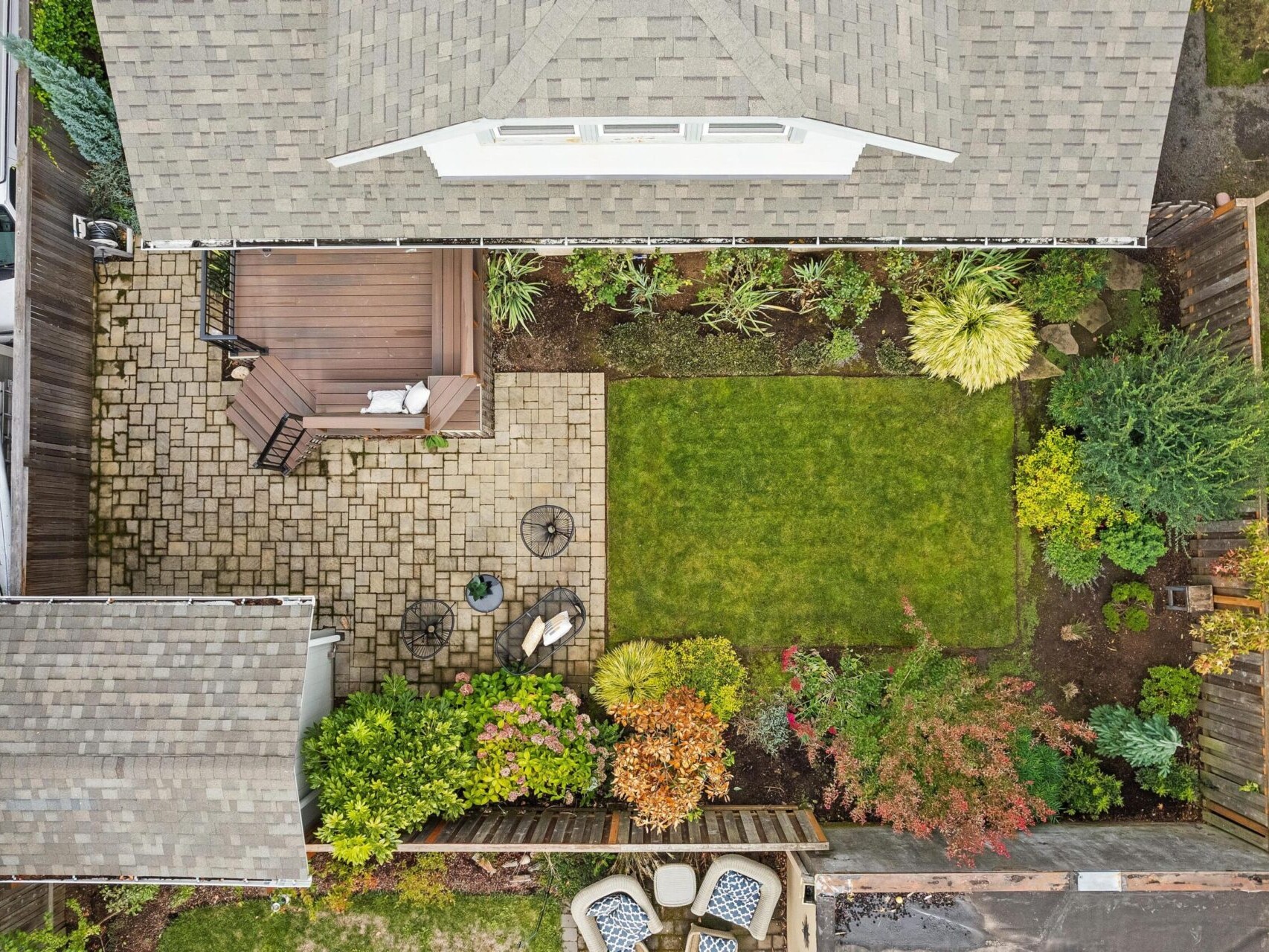 Aerial view of a backyard garden featuring a small lawn, a paved patio with seating, colorful plants along the edges, and two roofs partially visible. The space is enclosed by wooden fencing, creating a cozy and well-maintained outdoor area.