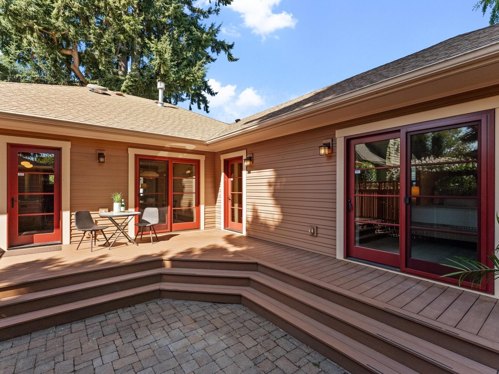 A spacious wooden deck with a small table and two chairs, nestled beside a tan Portland, Oregon house with red-framed patio doors. The sunlit area is beautifully complemented by tall trees visible in the background under a clear blue sky.