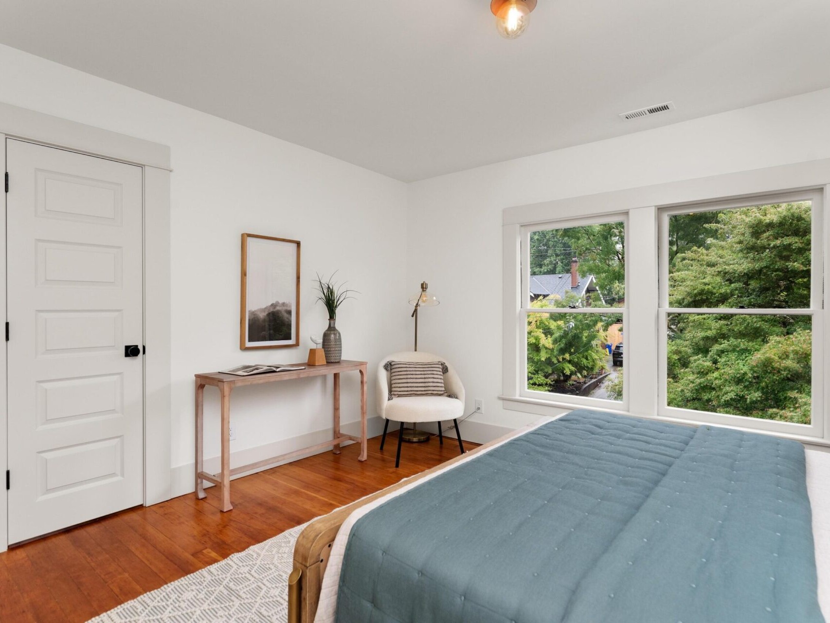 A cozy Portland, Oregon bedroom with a wooden floor, white walls, and a large window offering views of lush greenery. The room features a neatly made bed with a blue duvet, a small desk with decor items, and a chair adorned with a striped pillow.