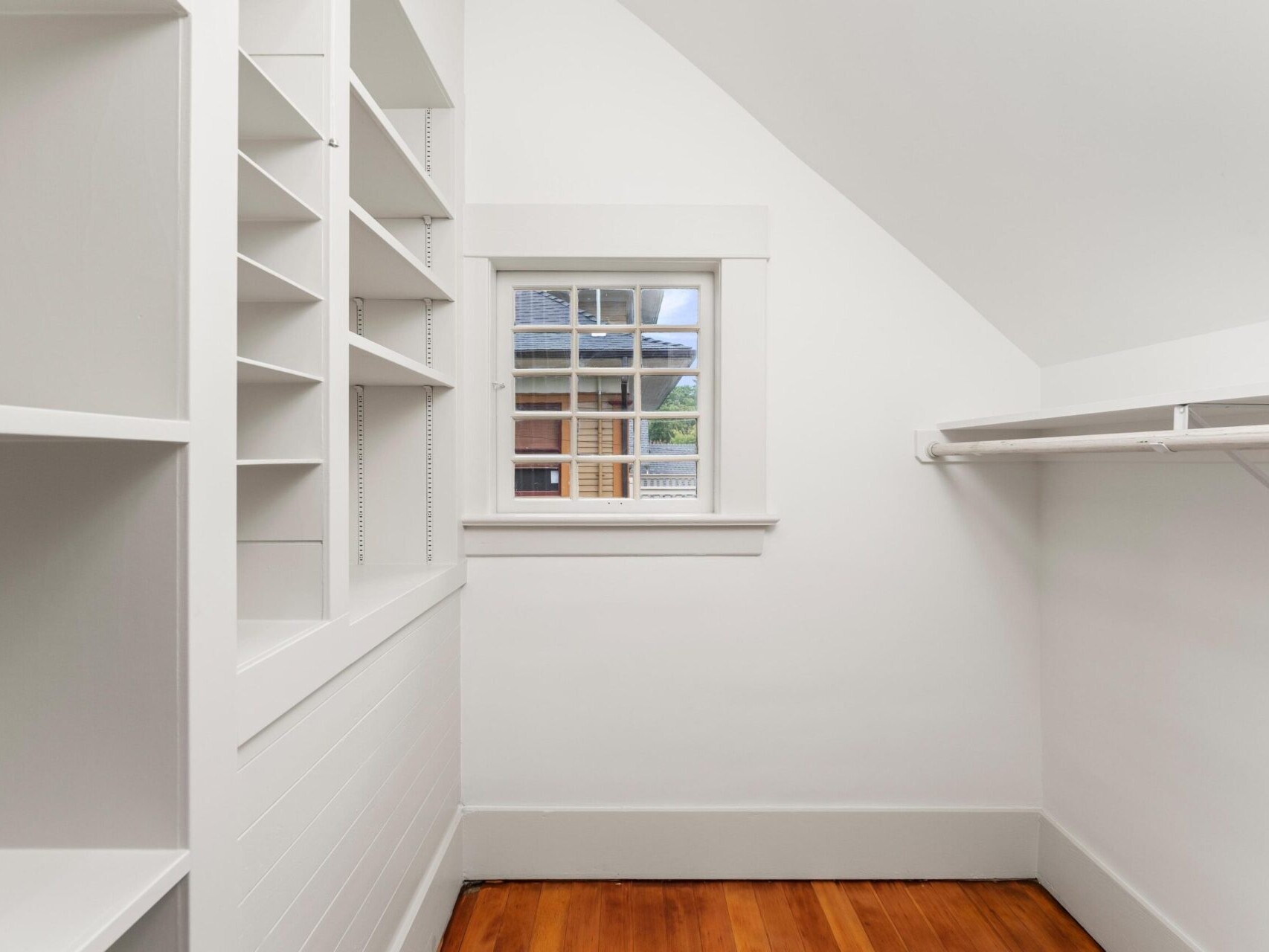 A small, white, empty walk-in closet with wooden floors awaits in Portland, Oregon. Built-in shelves line the left side, and an empty clothing rod hangs on the right. Near the back wall, a window offers a view of nearby buildings, capturing the city's unique charm.