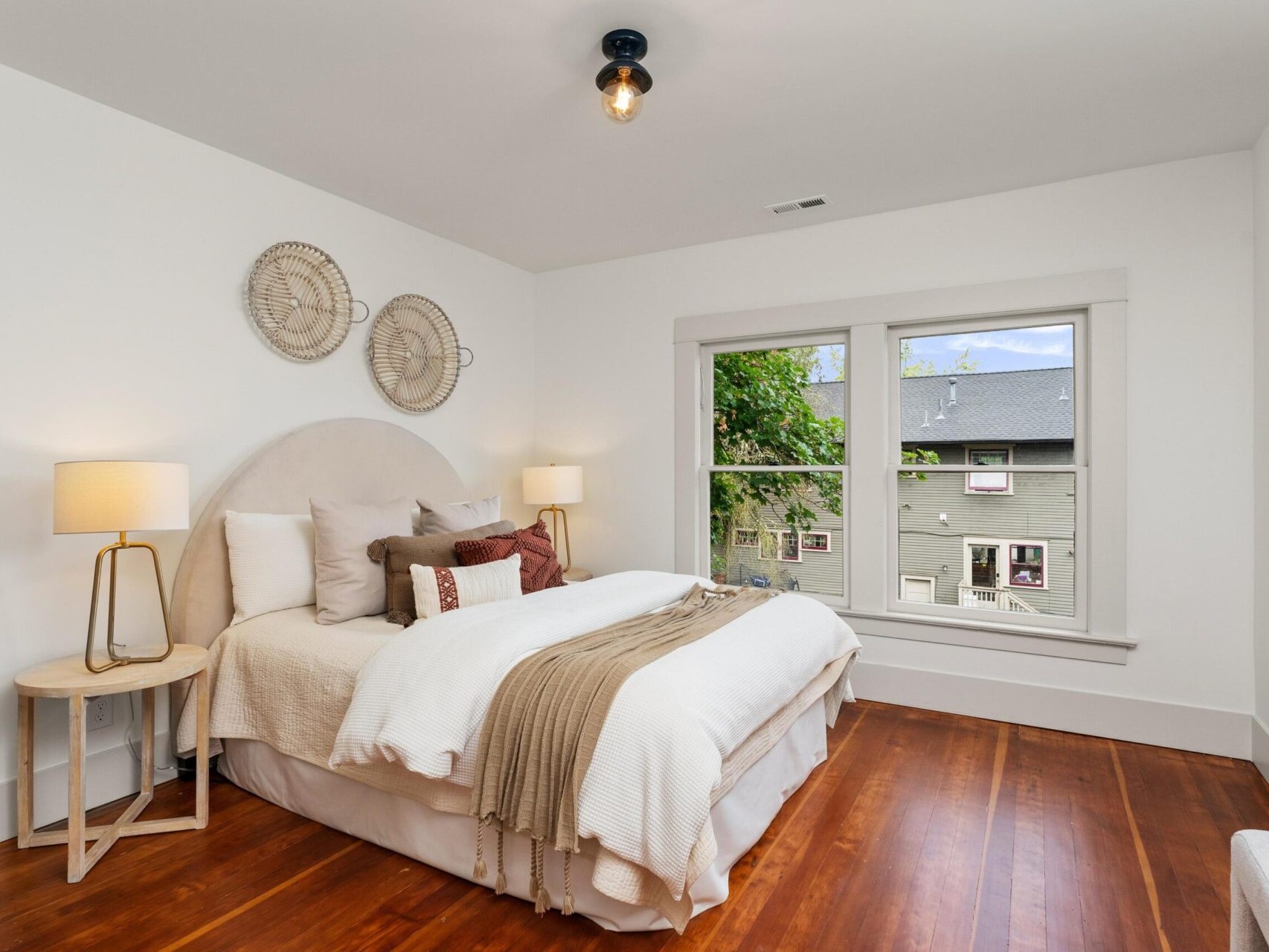 A cozy Portland, Oregon bedroom features a neatly made bed with white and beige bedding. Flanked by two side tables with lamps, the room boasts wooden floors and large windows showcasing a view of a neighboring house and trees. Elegant wall decor completes the inviting space.