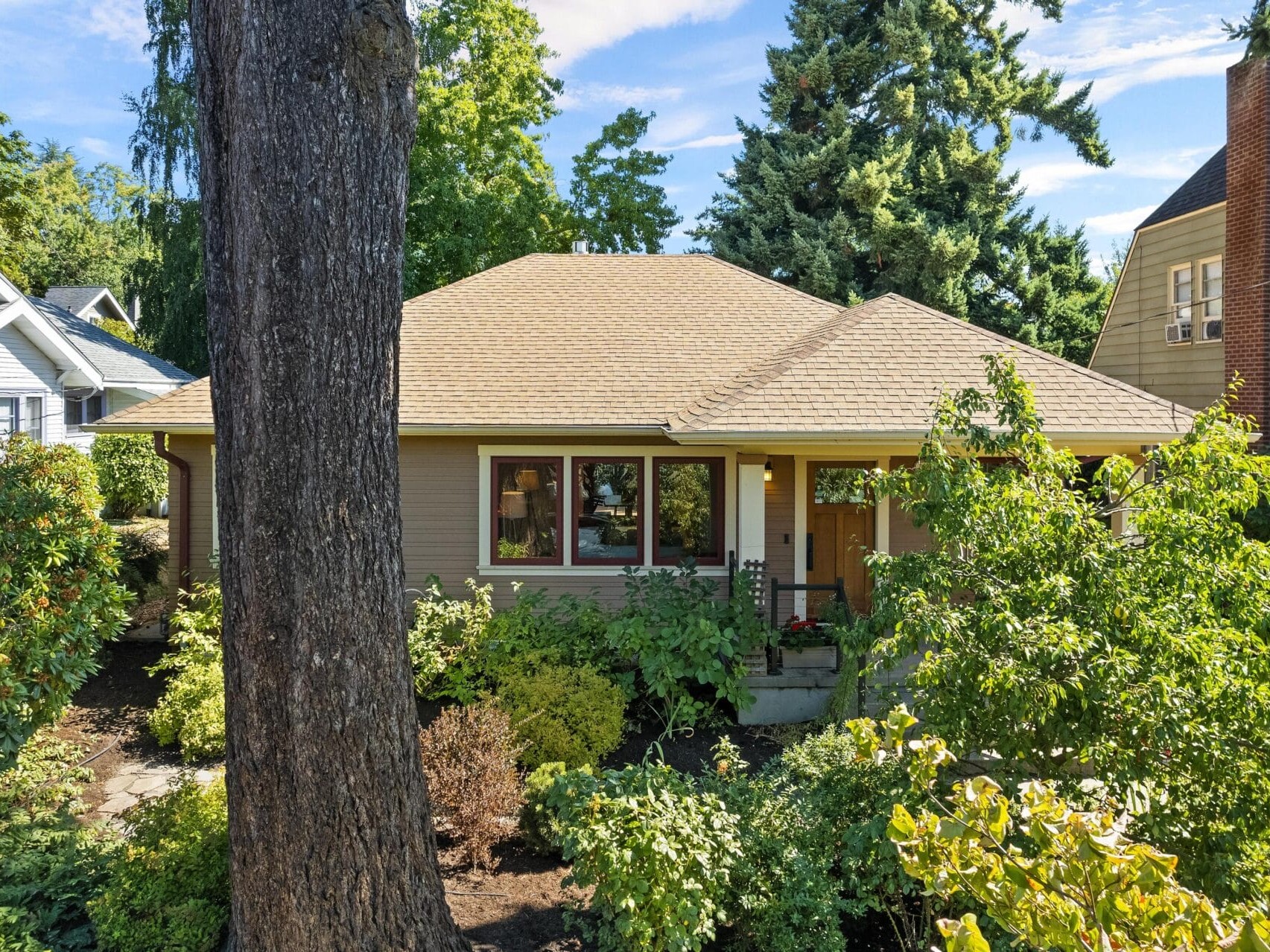 A cozy single-story house with a tan roof is nestled amidst Portland, Oregon's lush green trees and shrubs. The welcoming front entrance features a small porch, and the bright blue sky visible through the foliage adds a sunny, inviting atmosphere.