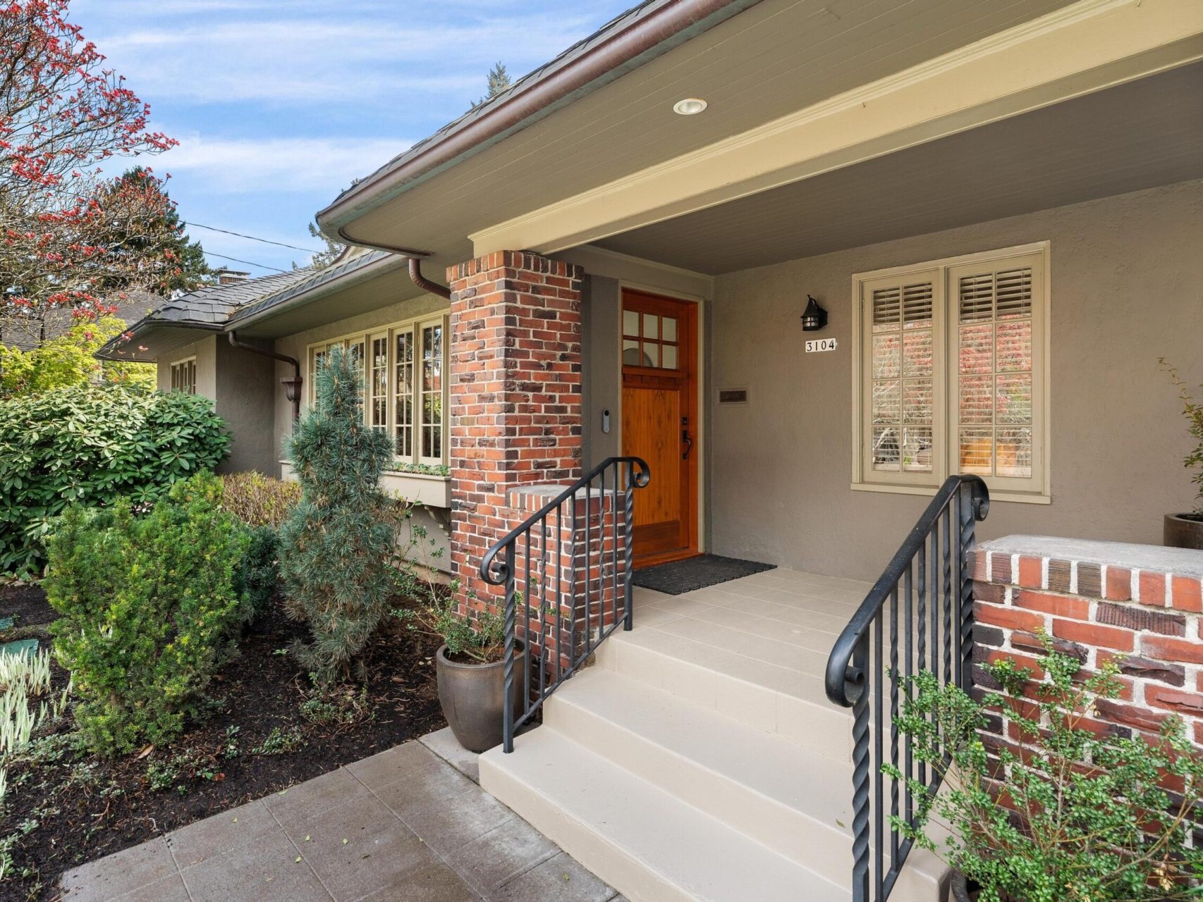 Front entrance of a Portland, Oregon real estate gem with red brick columns and beige siding. Steps lead to a wooden door, metal railings, and lush greenery including shrubs and trees under a partly cloudy sky, capturing the charm any Portland realtor would showcase.