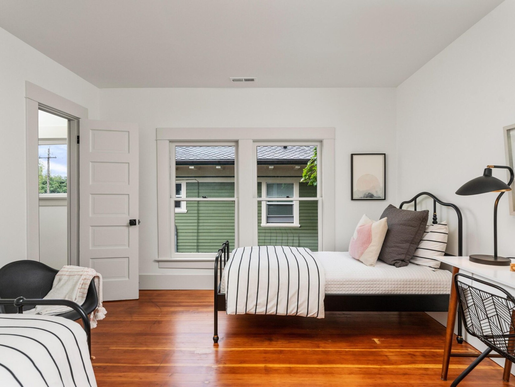 Minimalist bedroom in Portland, Oregon, with two single beds each adorned with striped bedding. Wooden floors lead to a window revealing an outdoor view of a green house. A black lamp and various decorations add charm to the space.