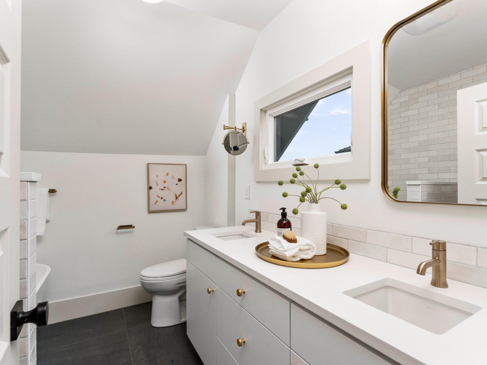 A modern Portland, Oregon bathroom features a double sink vanity with brass fixtures and two mirrors. Framed artwork adorns the wall above the toilet. A small tray with toiletries and a plant decorates the countertop, all beautifully illuminated by both natural and artificial light.
