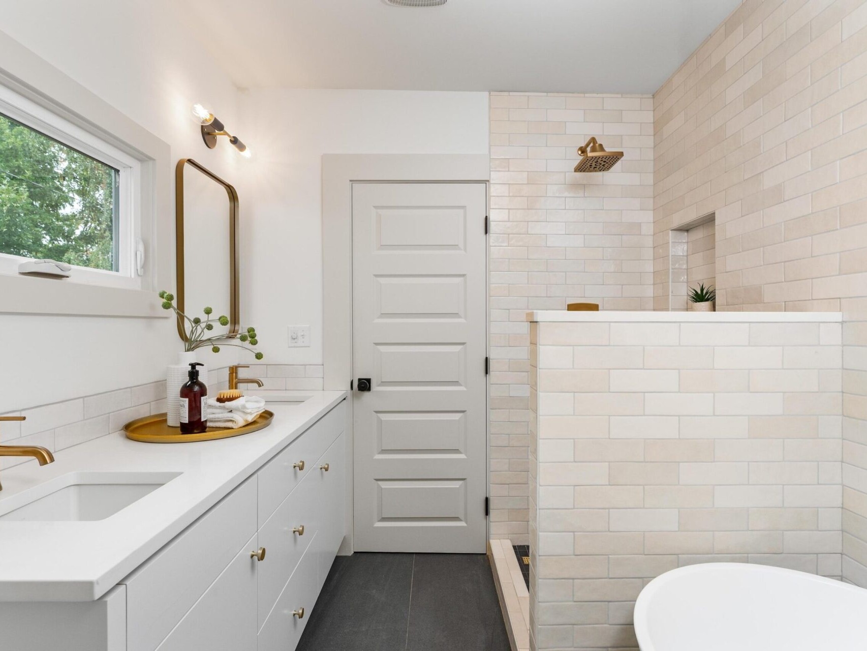A modern bathroom in a Portland, Oregon home features a white sink with gold faucet, a wooden tray with toiletries, and a rectangular mirror. A walk-in shower with beige tiles and gold fixtures is on the right, next to a standalone white bathtub. A small window graces the left wall.