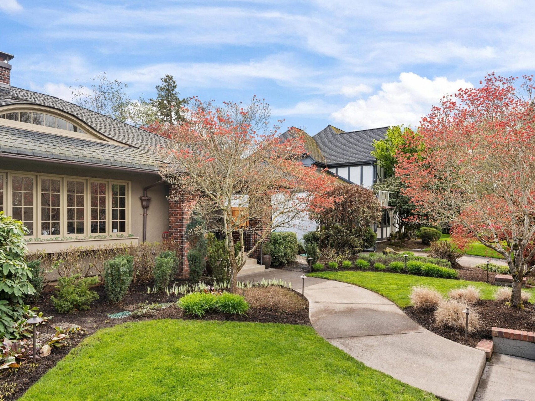 A charming suburban house in Portland Oregon with a landscaped front yard featuring a neatly trimmed lawn, small shrubs, and trees with red leaves. A curving concrete pathway leads to the entrance, set against a backdrop of a partly cloudy sky—ideal for exploration with your Portland Realtor.