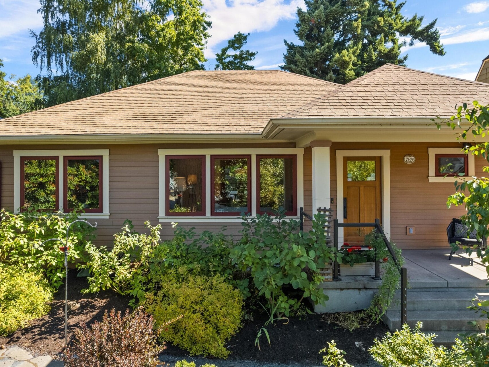 A single-story house in Portland, Oregon, boasts a tan exterior and a porch, surrounded by lush greenery and bushes. Four red-framed windows grace the front alongside a wooden door. The backdrop features tall trees reaching into the blue sky sprinkled with clouds.