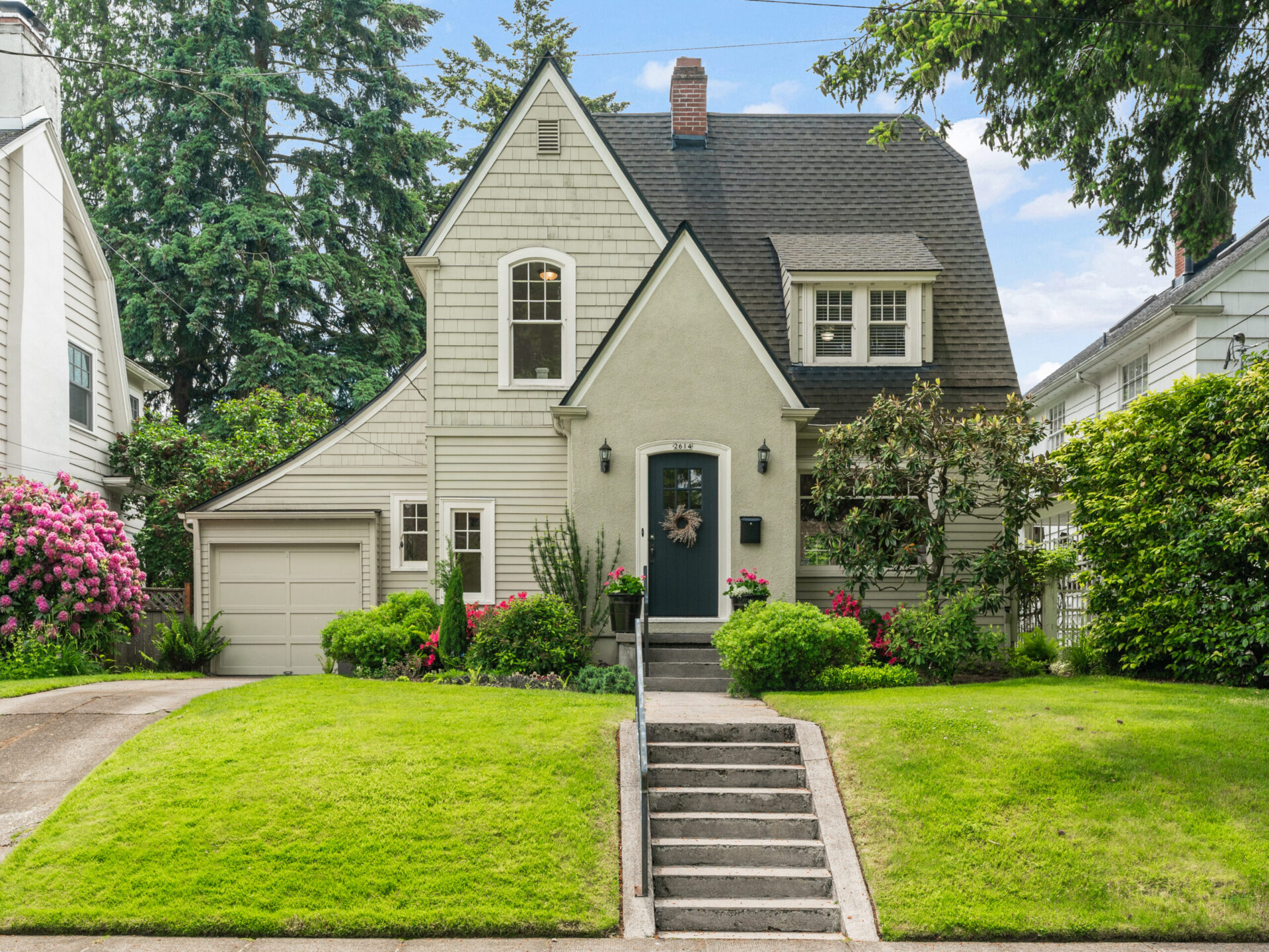A charming two-story house in Portland, Oregon, with a steeply pitched roof and light gray siding. The front yard boasts a manicured lawn, flower beds with pink flowers, and a stone walkway to the dark front door adorned with a wreath. Consult your Portland realtor to make this dream home yours.