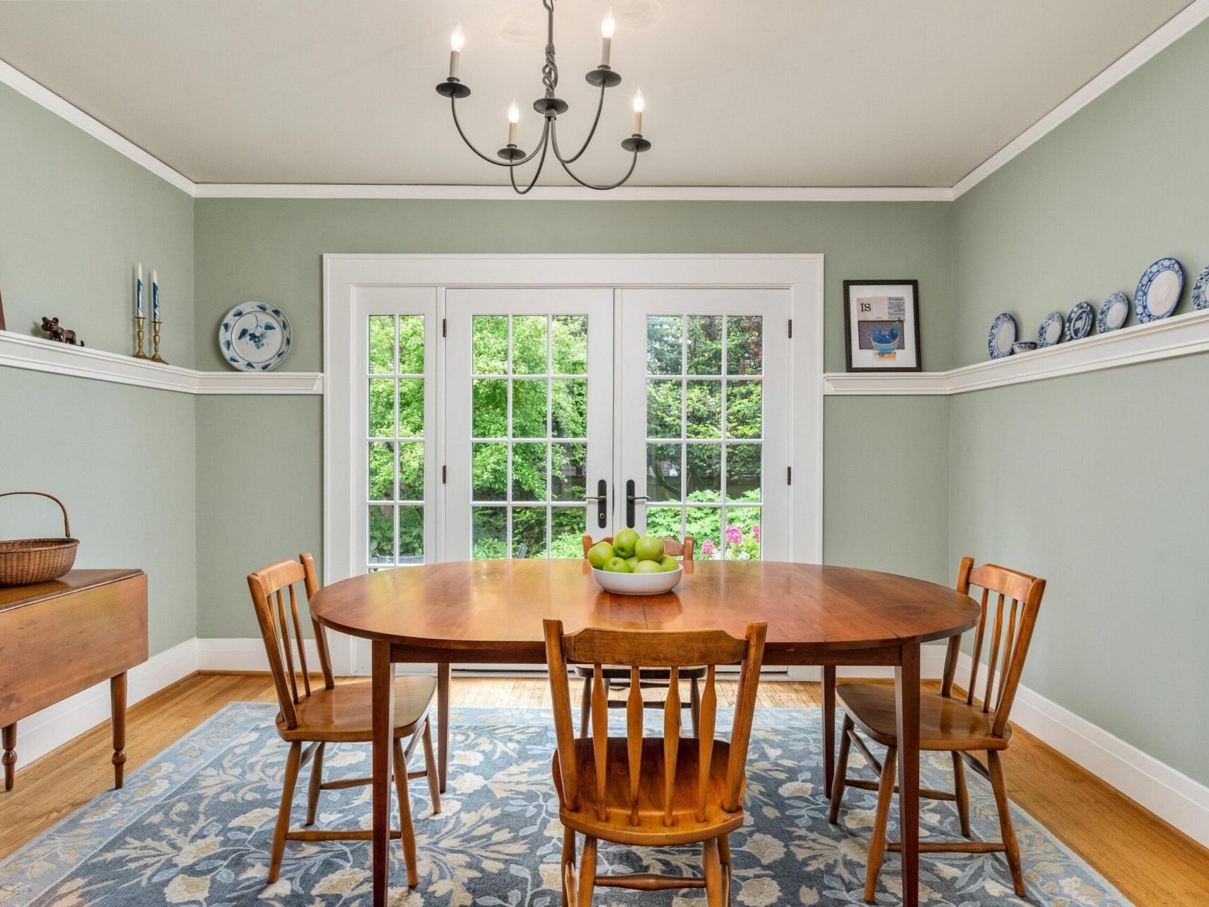 A dining room in Portland, Oregon, with light green walls features a wooden table and four chairs. A bowl of green apples sits on the table, while a side table holds a basket. Blue-patterned plates and art adorn the walls, and large windows reveal a garden view.