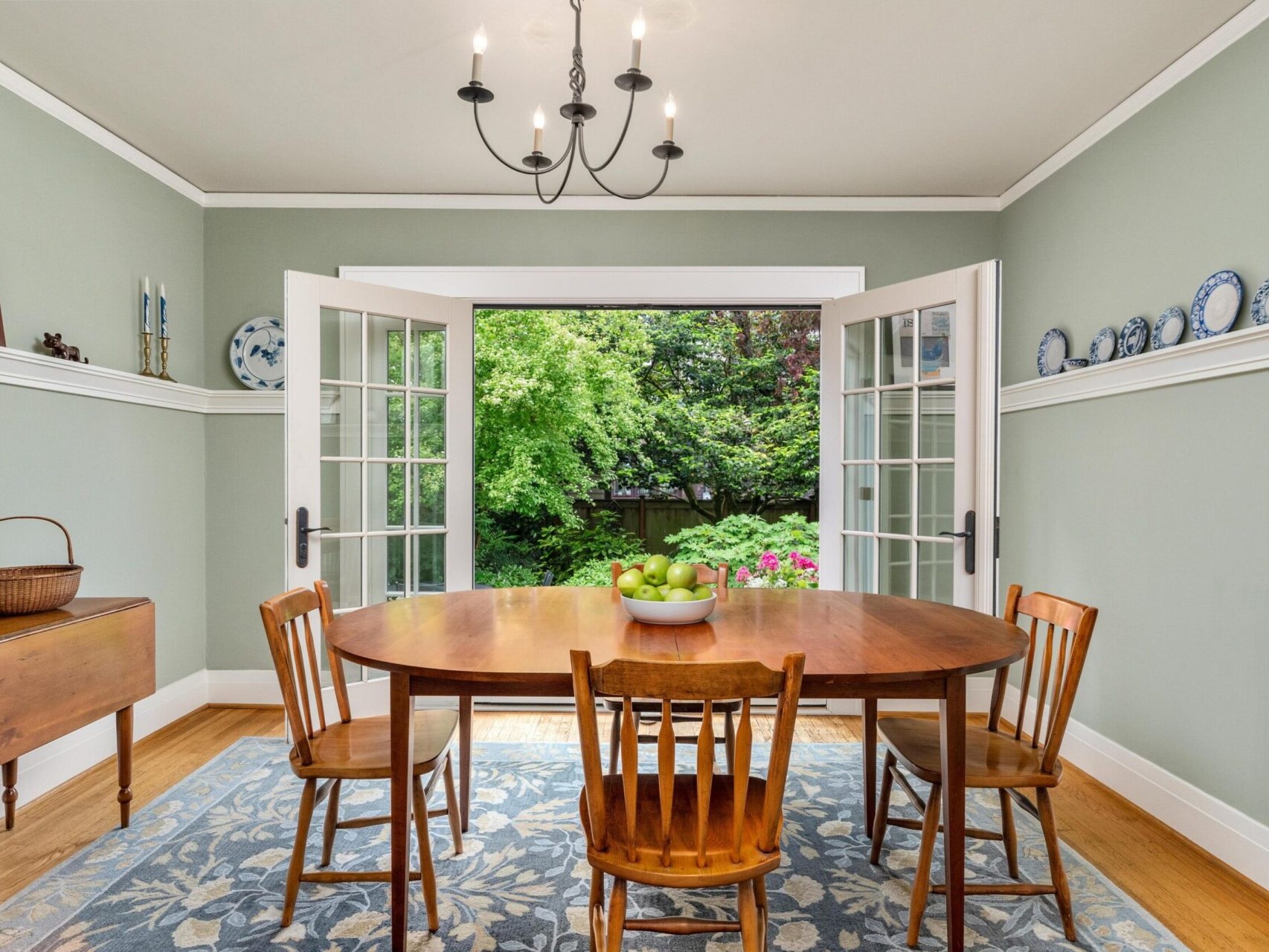 A dining room in Portland, Oregon features a wooden table and four chairs on a patterned rug. French doors open to a lush garden. A chandelier hangs from the ceiling, with decorative plates lining a shelf on the right wall. A basket and paintings adorn the left side.