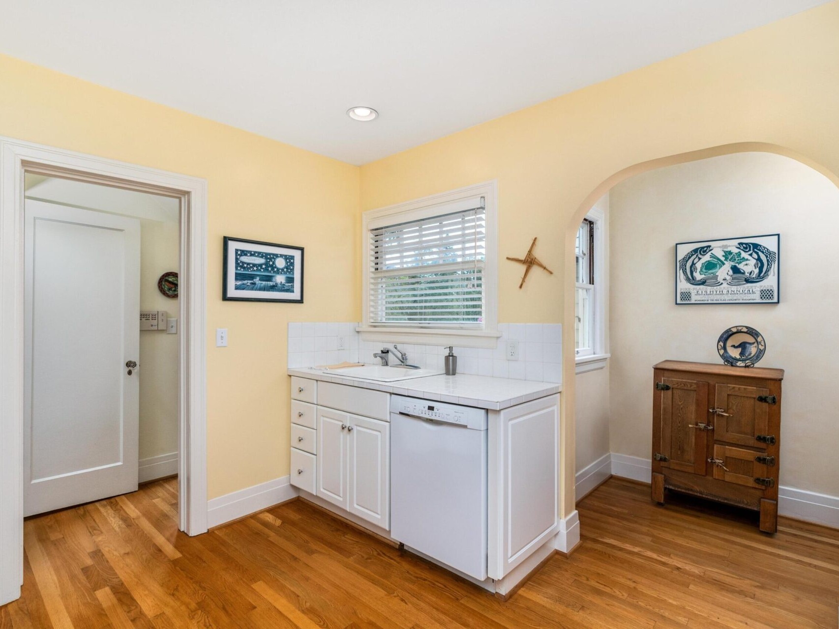 A cozy kitchen in Portland, Oregon, features yellow walls and wooden floors. There's a white dishwasher next to the sink and cabinets. A window with blinds is above the sink, while an archway leads to another room adorned with a wooden cabinet and nautical decor on the walls.