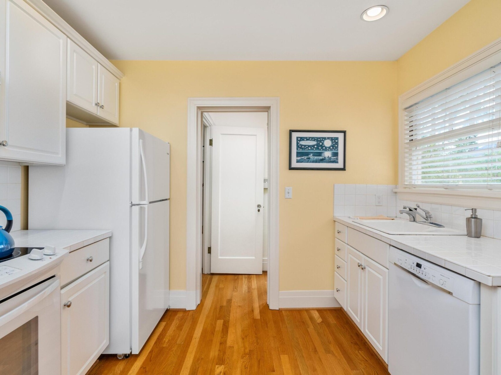 A bright kitchen in Portland, Oregon, boasts yellow walls and hardwood floors. It features white cabinets, a refrigerator, a dishwasher, and a stove with a blue kettle. A window with blinds sits above the sink while a framed picture adds charm to the wall.