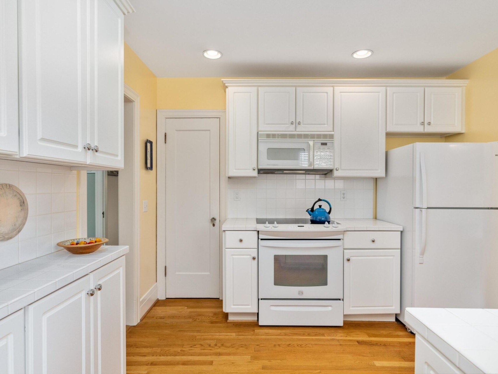 A bright kitchen with white cabinetry and appliances captures the charm of Portland, Oregon. The space features a refrigerator, stove, and microwave set against a yellow wall. A kettle sits on the stove, and a fruit bowl is placed on the wooden floor's counter.