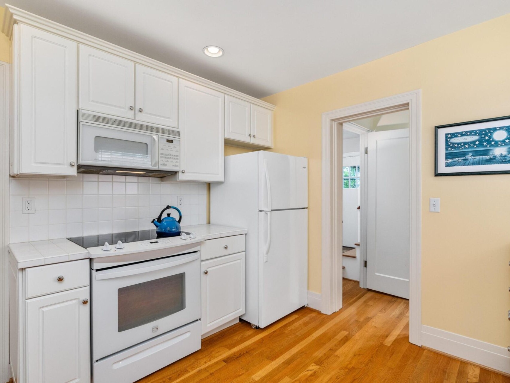 A cozy kitchen in Portland, Oregon features white cabinets and appliances, including a stove with a blue kettle. The pale yellow walls are adorned with a framed picture. The wooden floor adds warmth, and a doorway leads to another room.