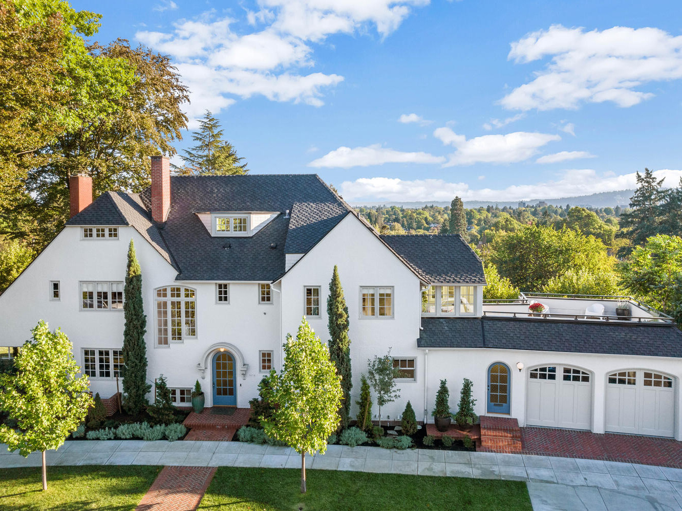 A large, elegant white house with a black roof set in the lush green landscape of Portland, Oregon. It features multiple windows, a brick driveway, and a two-car garage. Tall trees and a scenic view of distant hills enhance its picturesque setting.