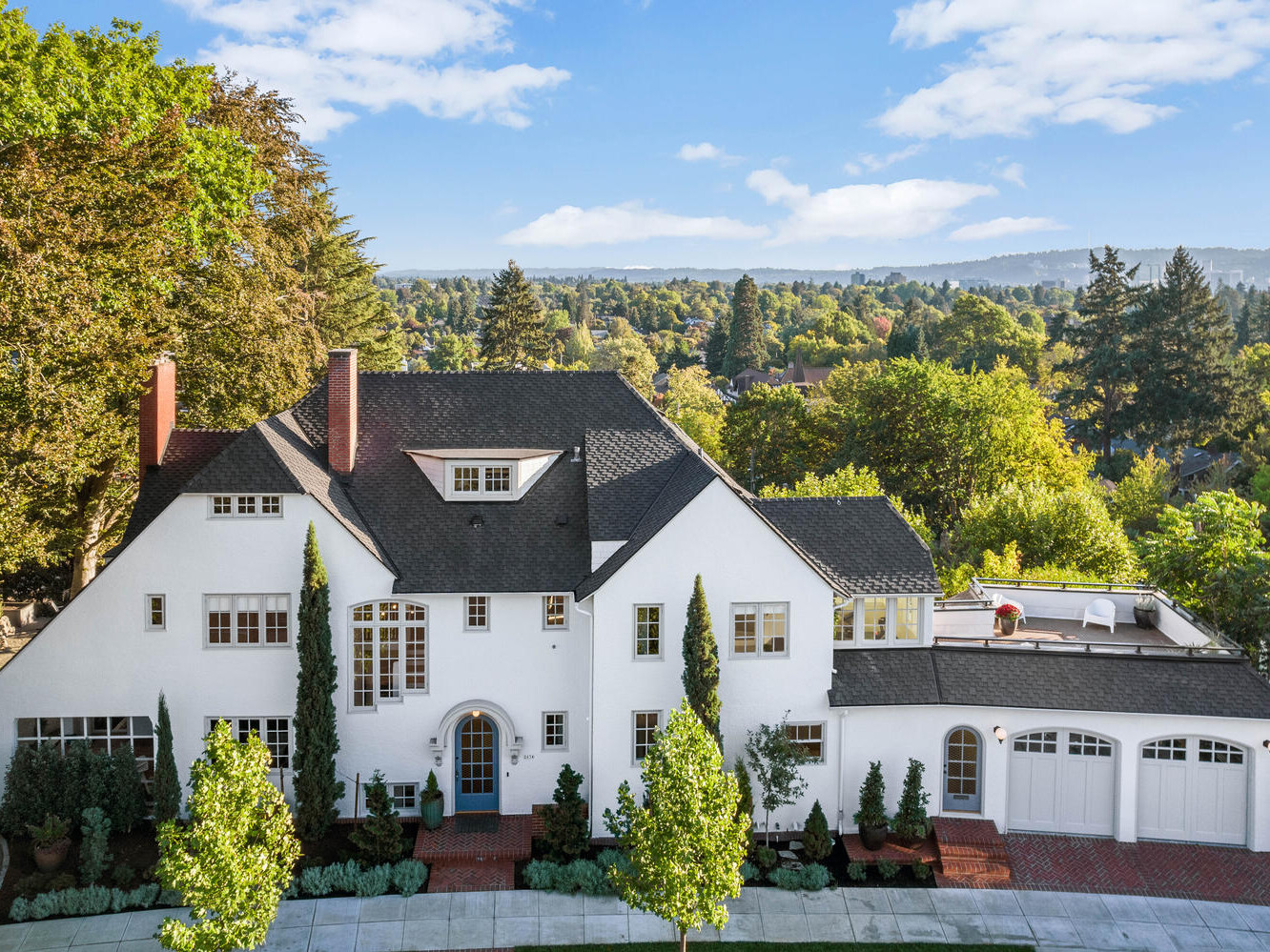 Aerial view of a large white two-story house with a black roof near Portland, Oregon, surrounded by lush greenery and trees. The house features multiple chimneys and a spacious patio area. The background reveals a scenic landscape with hills and a blue sky.