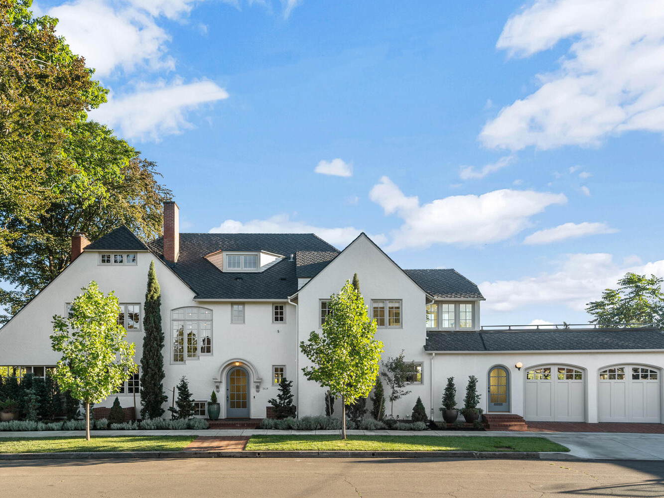 A large, two-story house in Portland, Oregon, with white stucco walls and a dark shingle roof is surrounded by greenery. It features an arched entrance, multiple chimneys, and an attached garage. The sky above is clear with a few clouds.