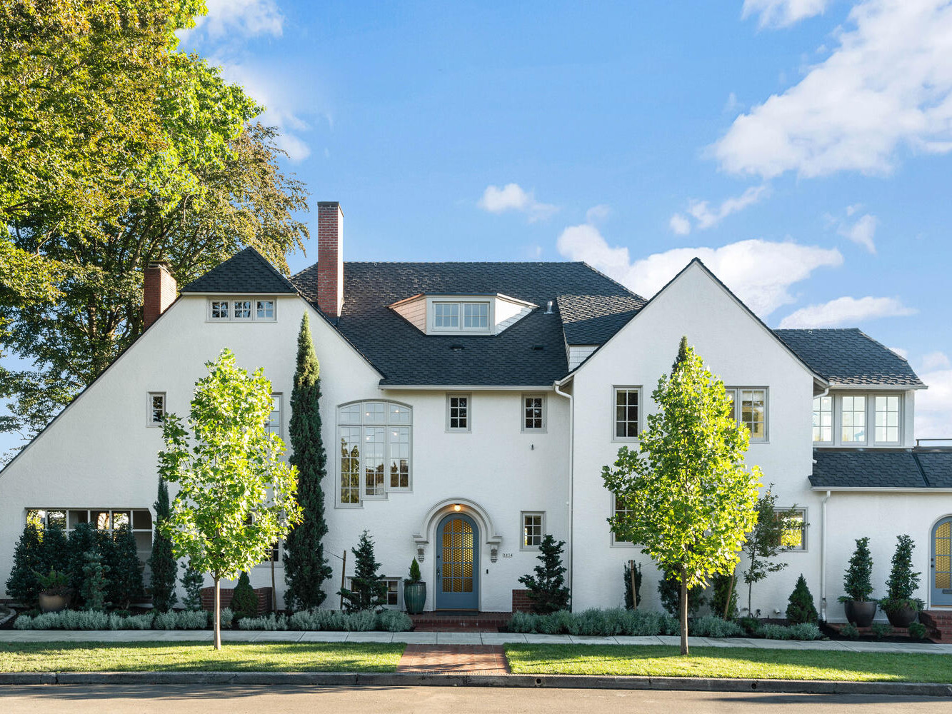 A large white house with a steep roof and multiple windows nestled in Portland, Oregon. Surrounded by trees and shrubs, the front yard features a red brick pathway and neatly trimmed greenery. The clear blue sky adds to its bright, inviting appearance.