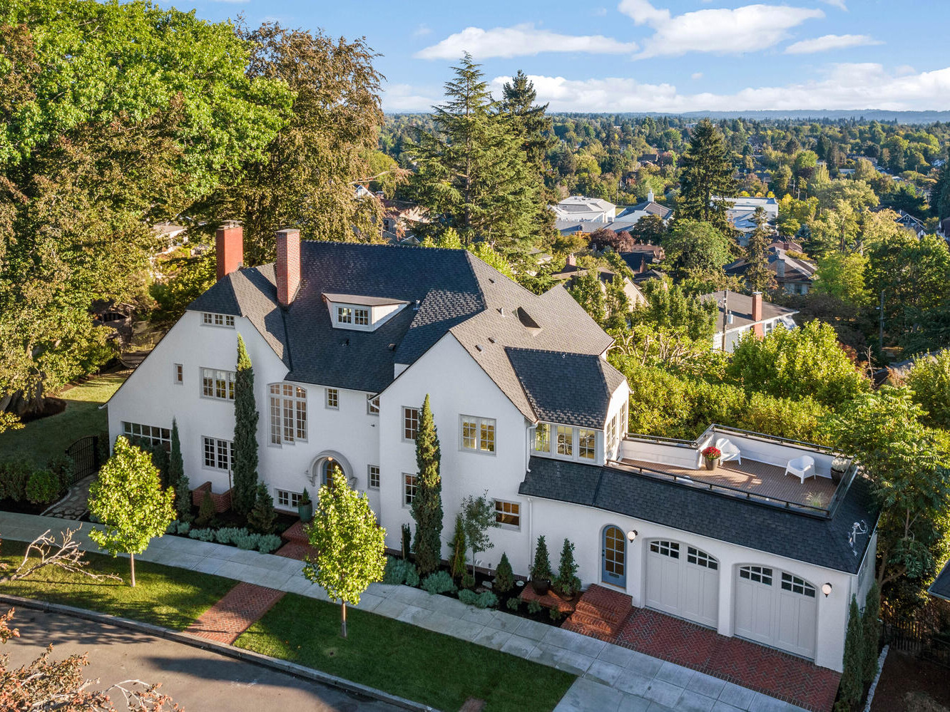 Aerial view of a large, white two-story house with a dark roof and several chimneys in Portland, Oregon. Surrounding trees provide greenery, and there's a patio area on the roof. The neighborhood features similar homes and is bathed in daylight.
