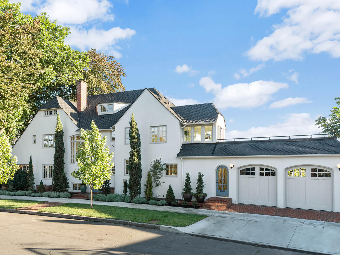 A charming white house with a black roof in Portland, Oregon, is surrounded by tall trees and shrubs. It features large windows, a double garage with arched doors, and a brick driveway. The blue sky above is dotted with scattered clouds.