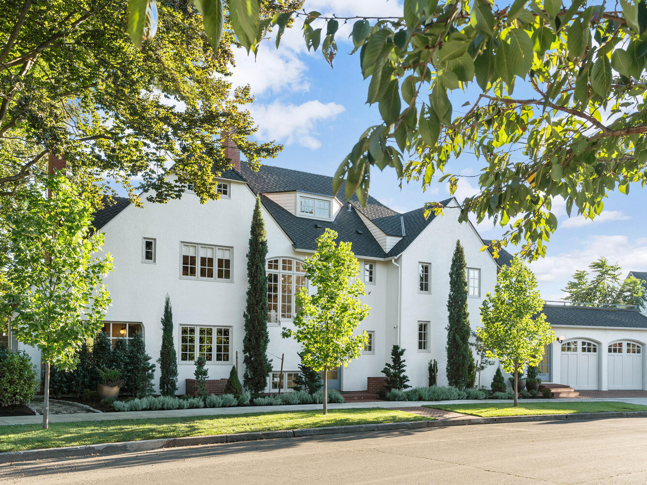A large white house with a black roof surrounded by Portland, Oregon's lush greenery and trees. The facade features multiple tall windows and two garage doors. The front yard is neatly landscaped, and the scene is lit by bright, clear daylight.