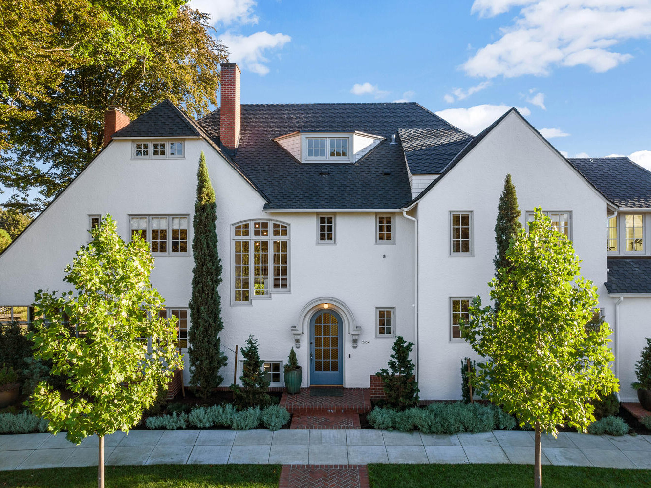 Charming two-story white house in Portland, Oregon with a steeply pitched roof, surrounded by lush greenery and tall trees. Featuring large arched windows, a brick pathway, and symmetrical landscaping under a bright blue sky with scattered clouds.