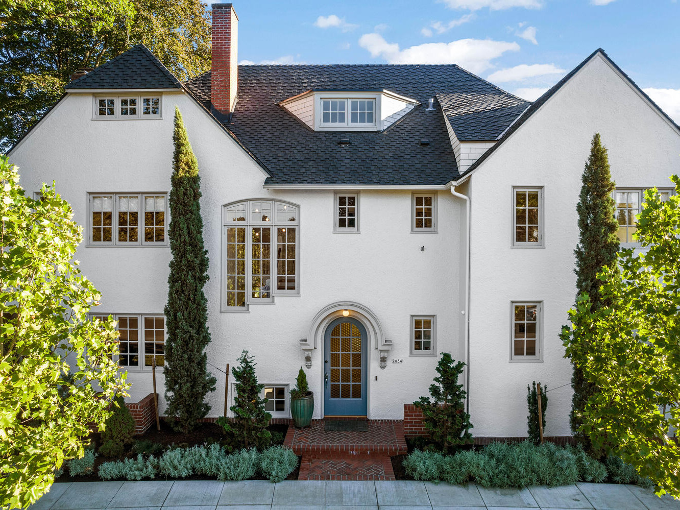 A large, elegant white stucco house in Portland, Oregon boasts steep dark roofs and multiple windows. Tall trees frame the entrance, which features an arched doorway and decorative plants. The sky is clear and blue.