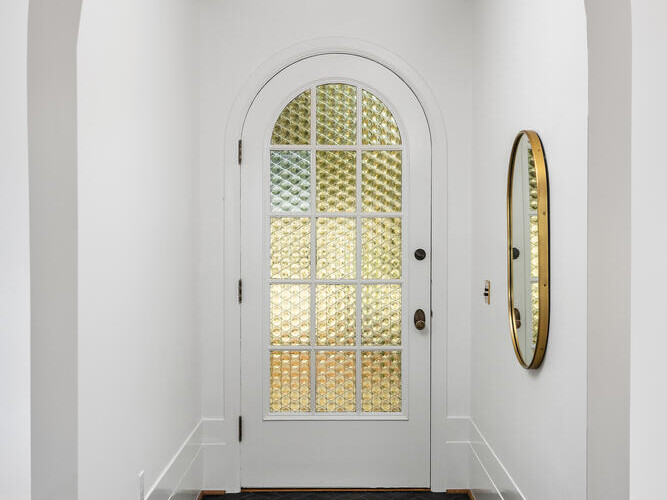 A narrow hallway with white walls evokes a chic Portland, Oregon, vibe, featuring a glass-paneled arched door at the end. A round mirror hangs on the right wall. The floor boasts a dark herringbone pattern, while a small ceiling light fixture illuminates the space.
