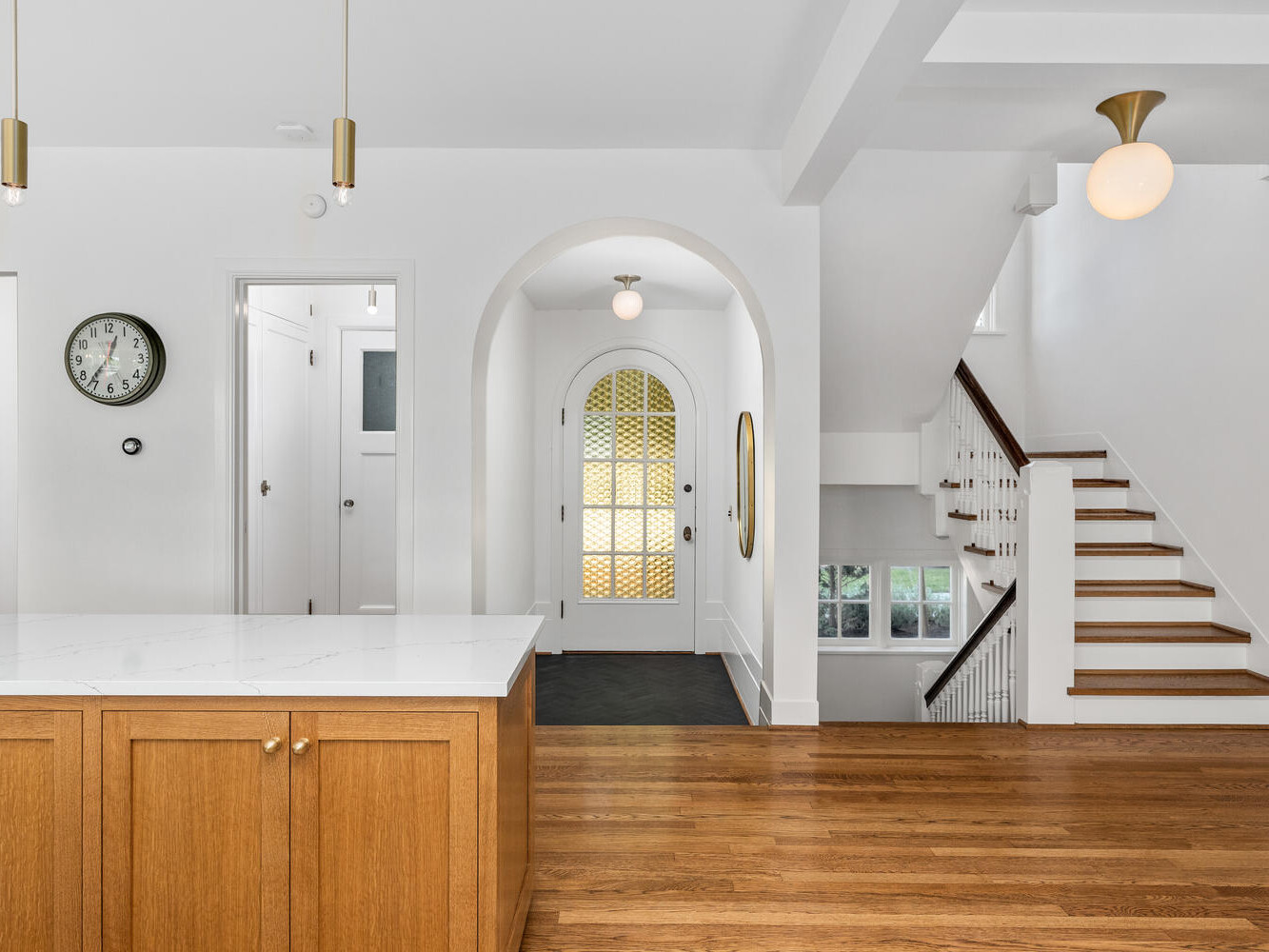 Bright kitchen with wooden floors, a white marble island, and gold-accented pendant lights. An arched doorway leads to a hallway with a yellow-patterned glass door. A staircase with wooden steps ascends to the right. In Portland, Oregon, a wall clock reads 10:10.