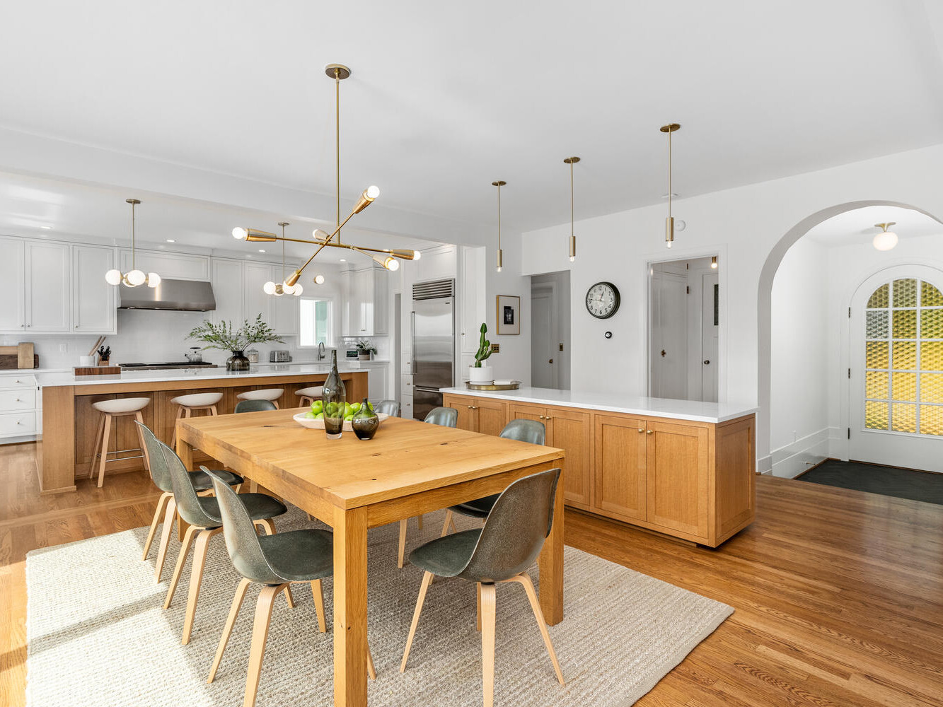 A modern kitchen and dining area in Portland, Oregon, with wooden floors and furniture. It features a large wooden dining table with green chairs, a kitchen island with bar stools, pendant lighting, and white cabinetry. A round archway reveals an adjoining room.