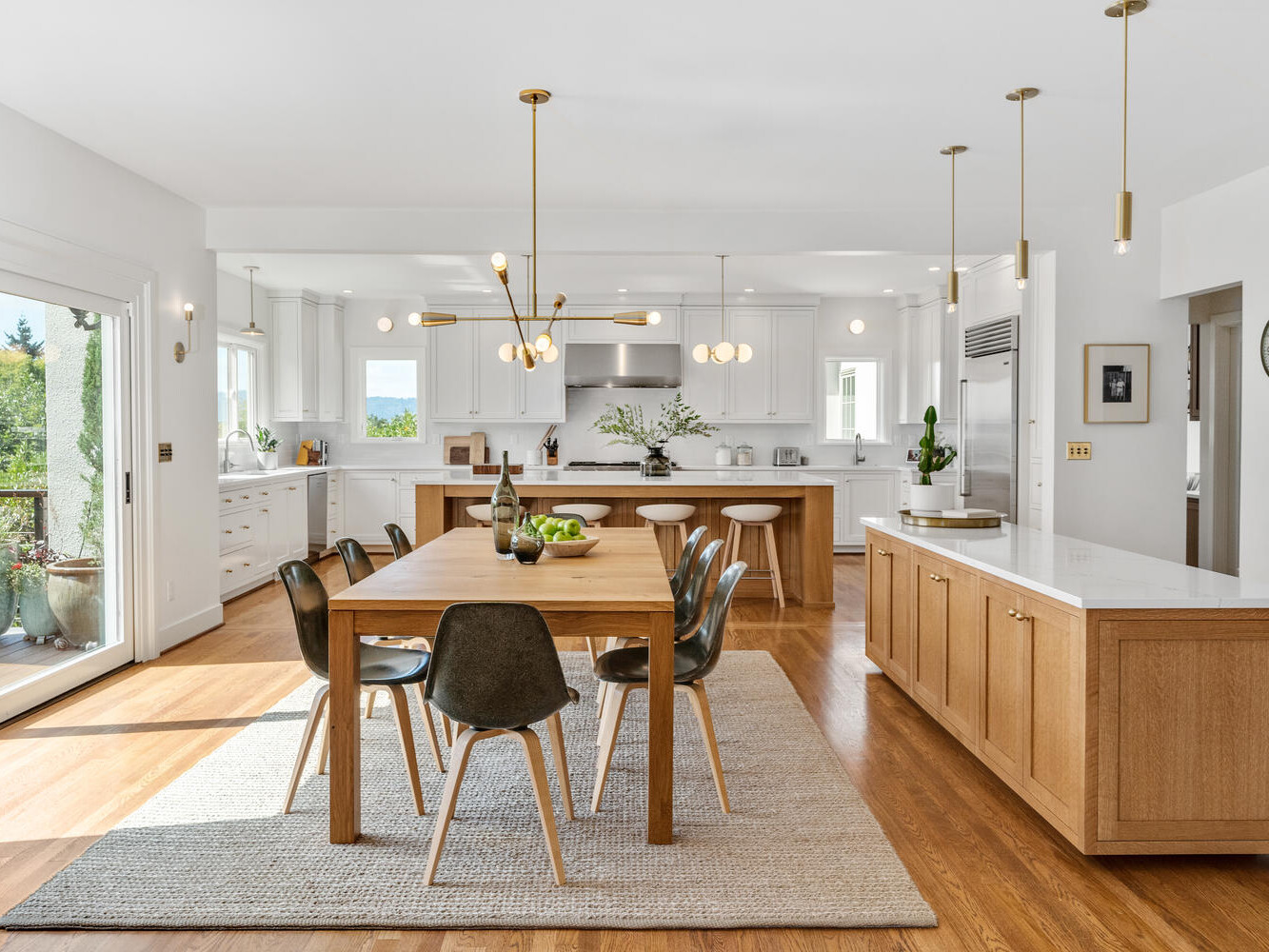 A spacious, modern kitchen and dining area in Portland, Oregon features wooden floors and sleek white cabinets. A wooden dining table with black chairs sits on a rug. Pendant lights hang above, while large windows flood the space with natural light. A wall clock and plants add charm.