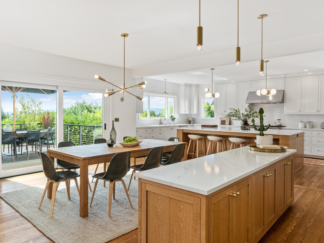 Spacious modern kitchen with white cabinetry, marble countertops, and wooden accents. An island with stools centers the room, while a dining table with chairs is nearby. Floor-to-ceiling windows provide natural light and a view of a patio and Portland Oregon's lush greenery outside.