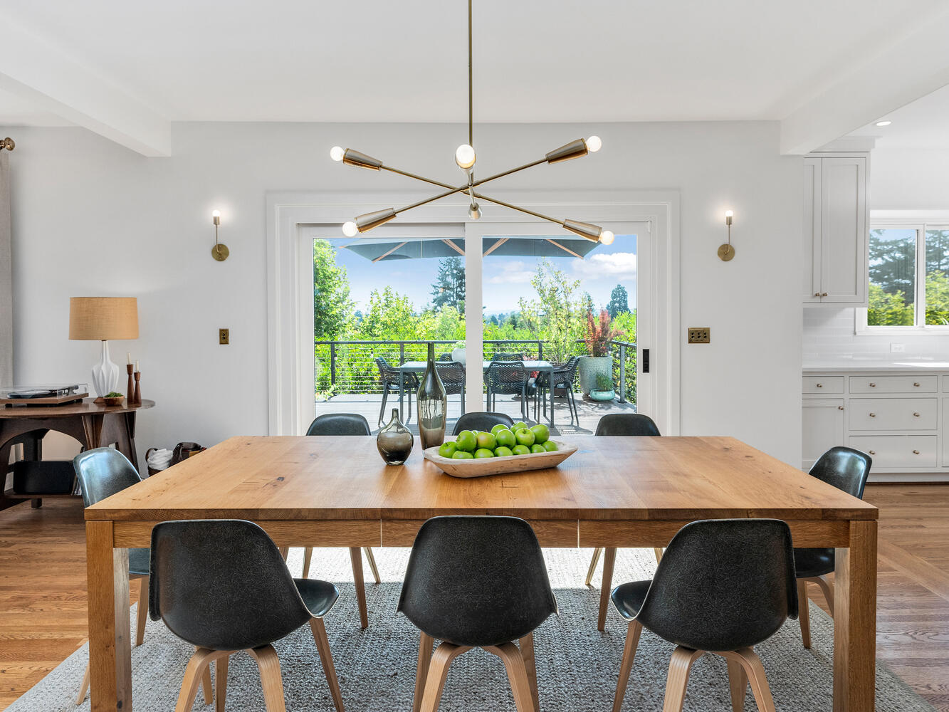 A bright dining room in Portland, Oregon, features a wooden table with a bowl of green apples and black chairs. A modern chandelier hangs above. Large glass doors open to a patio with views of trees and open skies. White walls and hardwood floors complete the space.