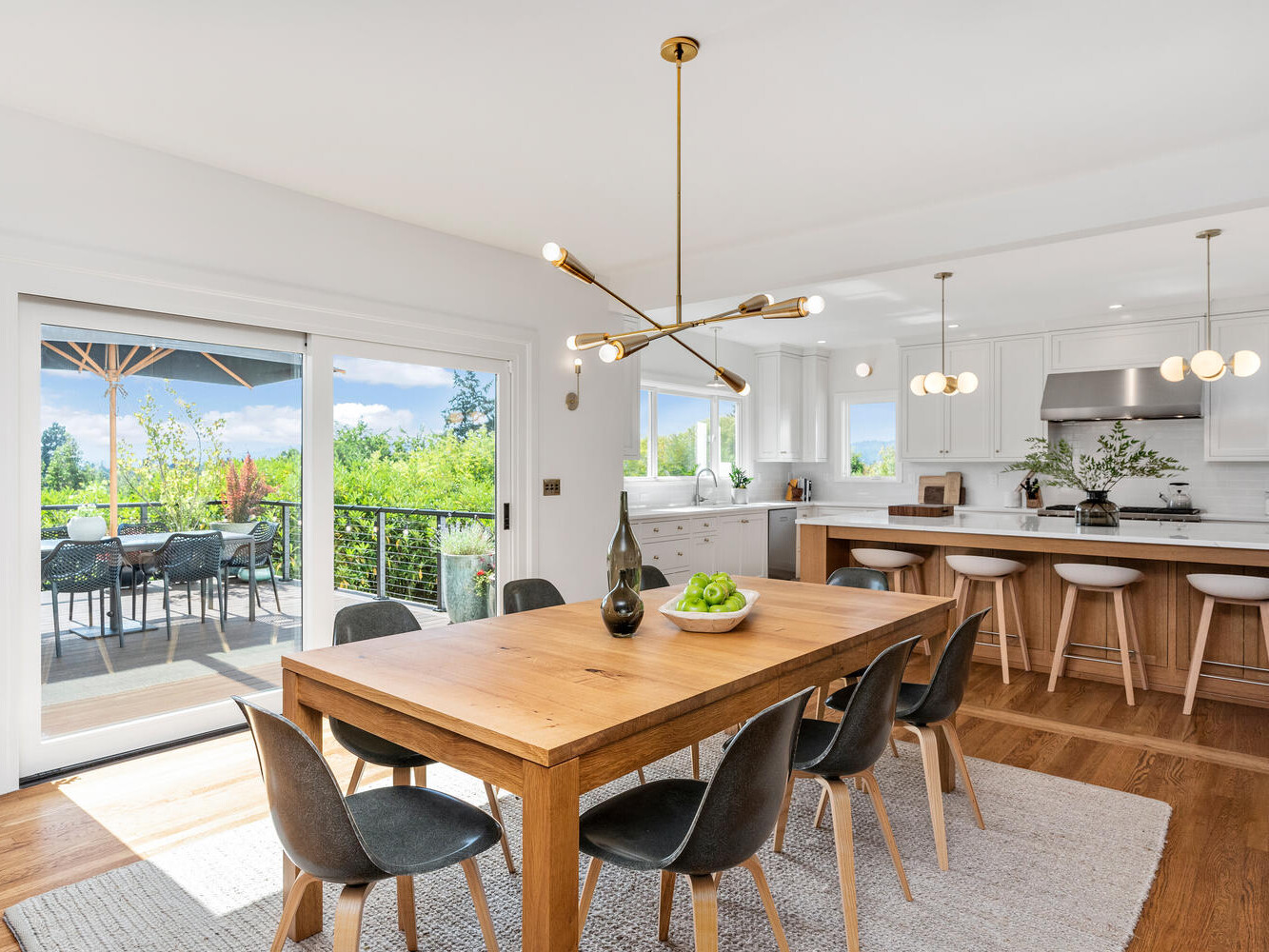 A bright, modern kitchen and dining area in Portland, Oregon, features a large wooden table with black chairs beneath a unique chandelier. Sliding glass doors reveal an outdoor patio and lush greenery. The kitchen boasts white cabinets and a spacious island with bar stools.