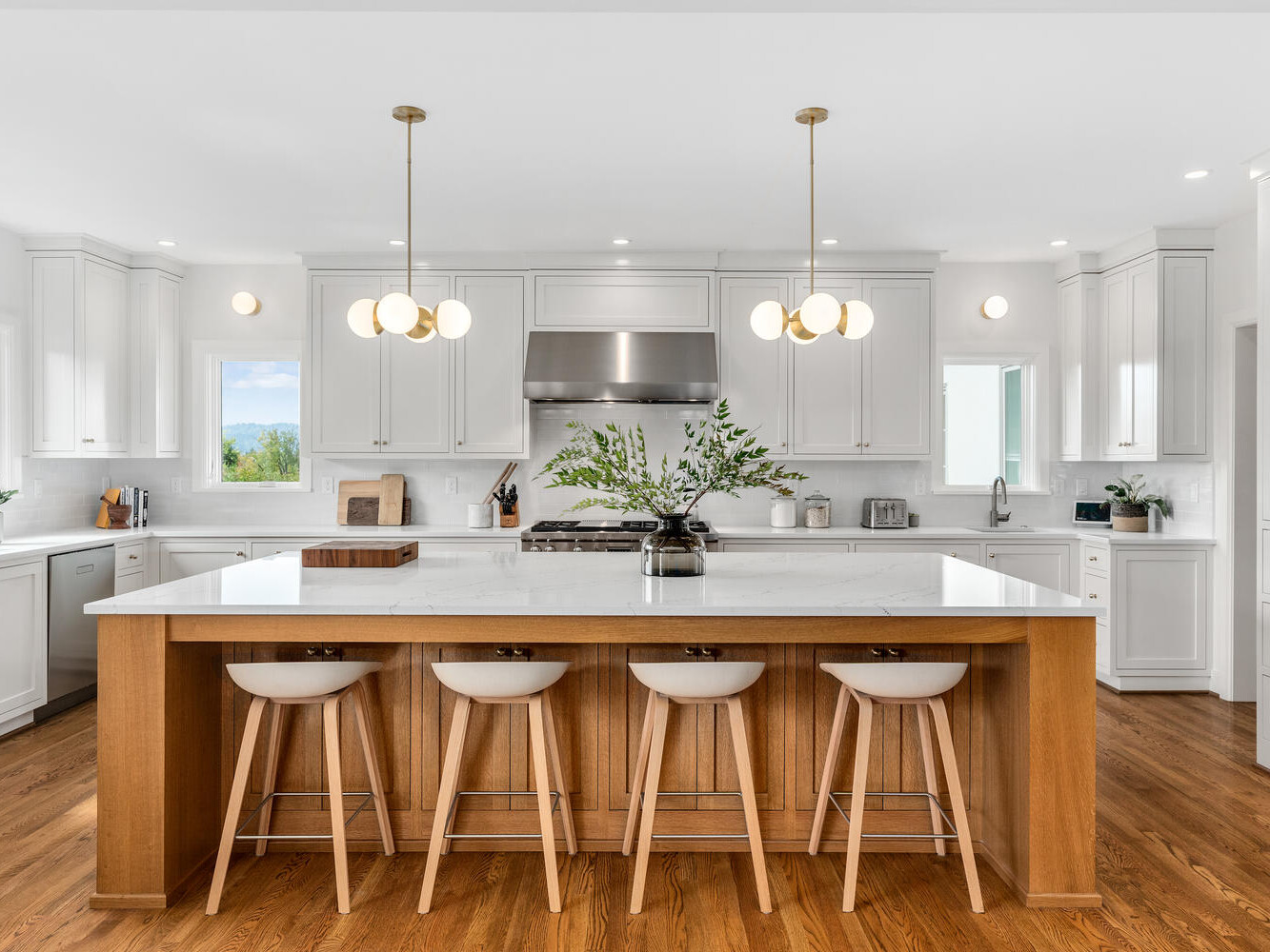 A spacious modern kitchen in Portland, Oregon features white cabinetry and a large wooden island. Four white stools align in front, and three globe pendant lights hang above. Stainless steel appliances and a vase with greenery add accents. Natural light fills the room.