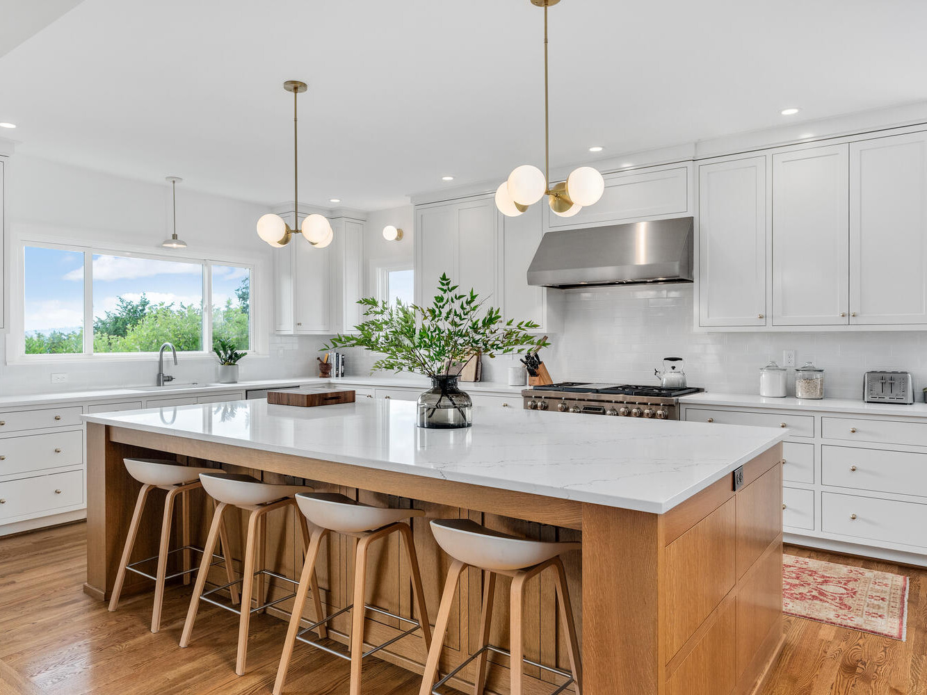 A modern kitchen in Portland, Oregon features a large island with a white marble countertop and wooden base, accompanied by four bar stools. Above are three pendant lights. The space boasts white cabinets, stainless steel appliances, and a window offering views of lush trees.