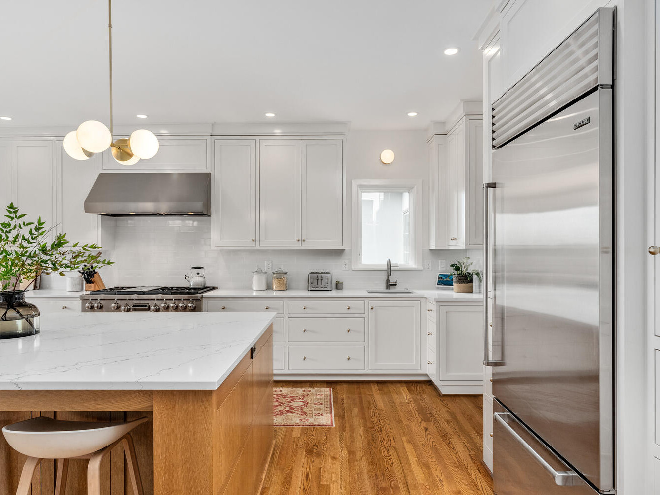 A modern kitchen in Portland, Oregon, boasts white cabinetry and a stainless steel fridge. A large island with a marble countertop and wooden stools takes center stage. A plant and pendant lights add decoration, while hardwood floors enhance the warm ambiance.