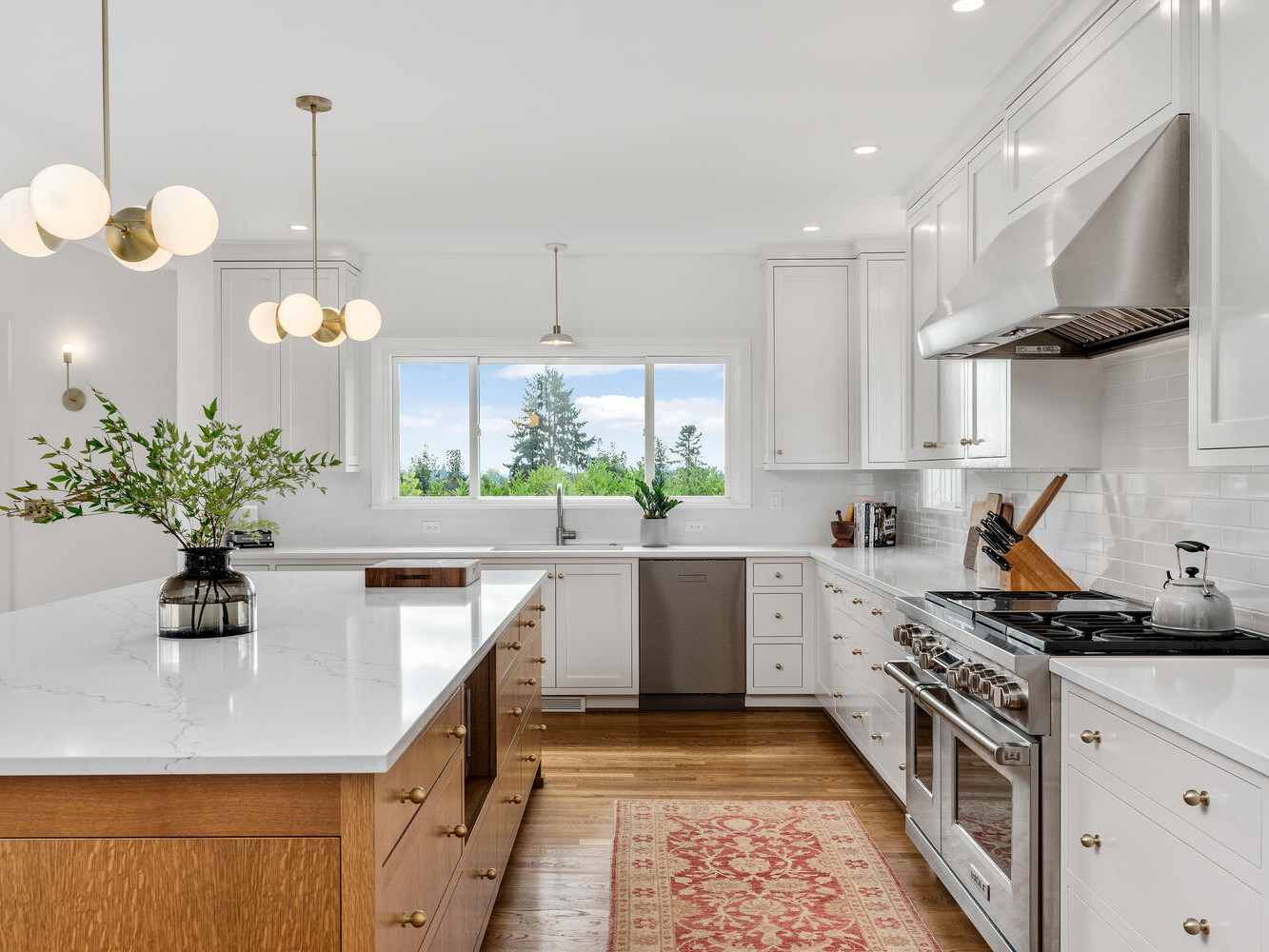 Bright, modern kitchen in Portland, Oregon, with white cabinets and marble countertops. Central island features a wooden base and stylish pendant lights. Stainless steel appliances and a view of greenery through a large window complete the sleek look.